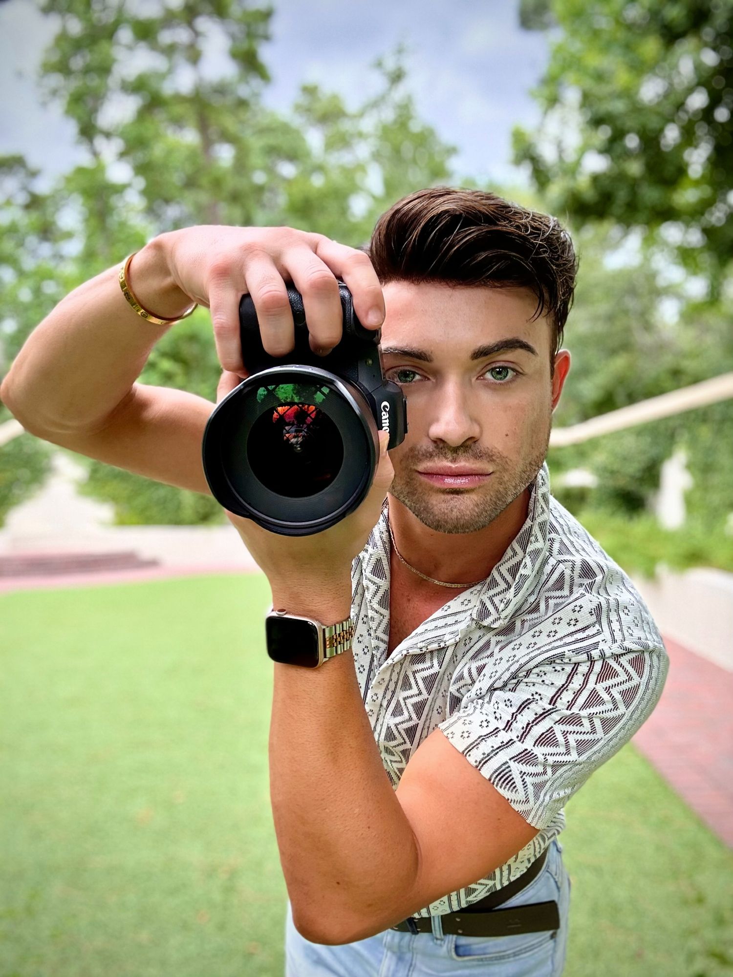 A photographer holds a professional camera with a wide-angle lens outdoors while wearing a patterned short-sleeve shirt.