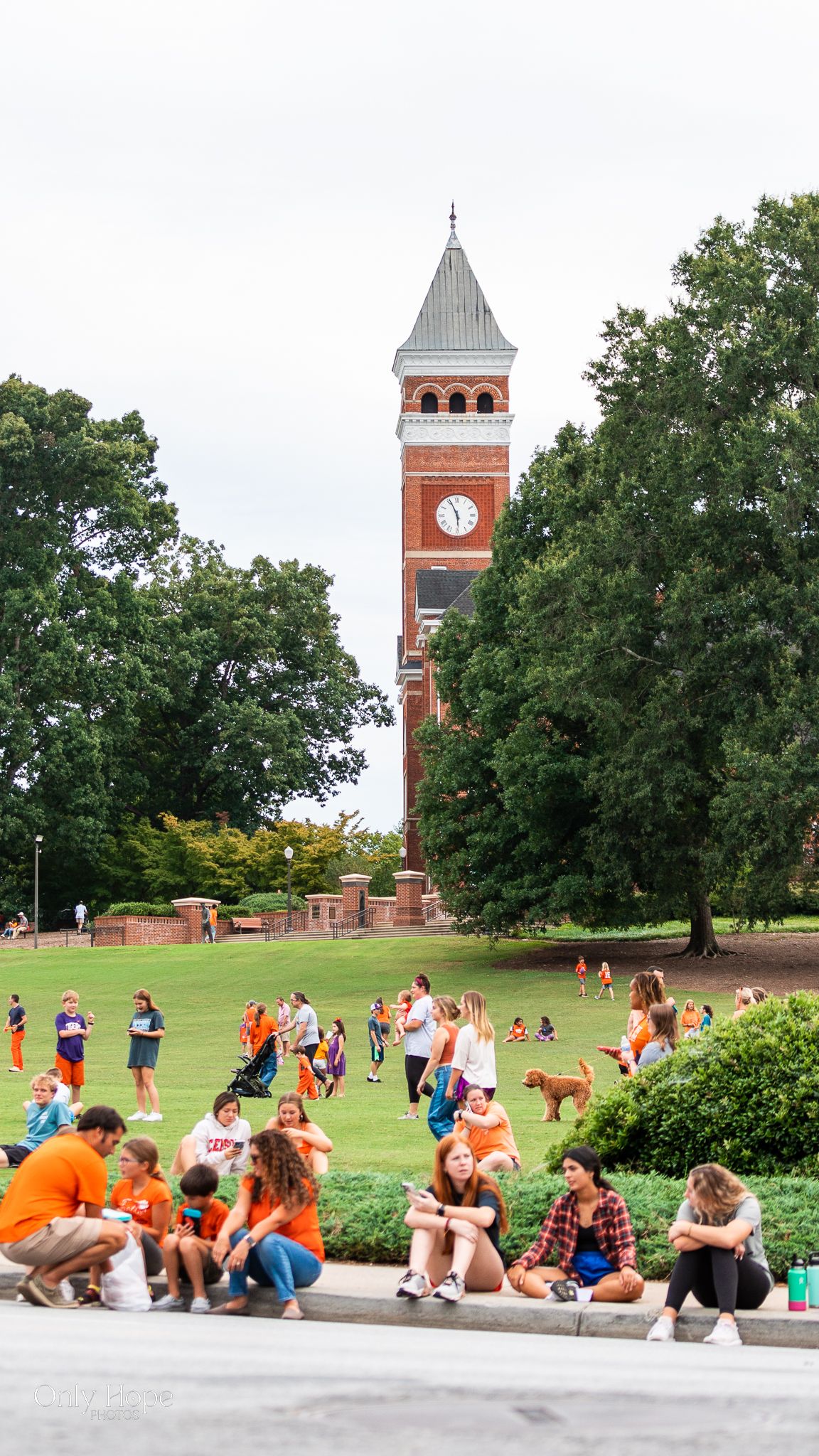 Students gather on a university lawn beneath a tall brick clock tower surrounded by trees on a sunny day.