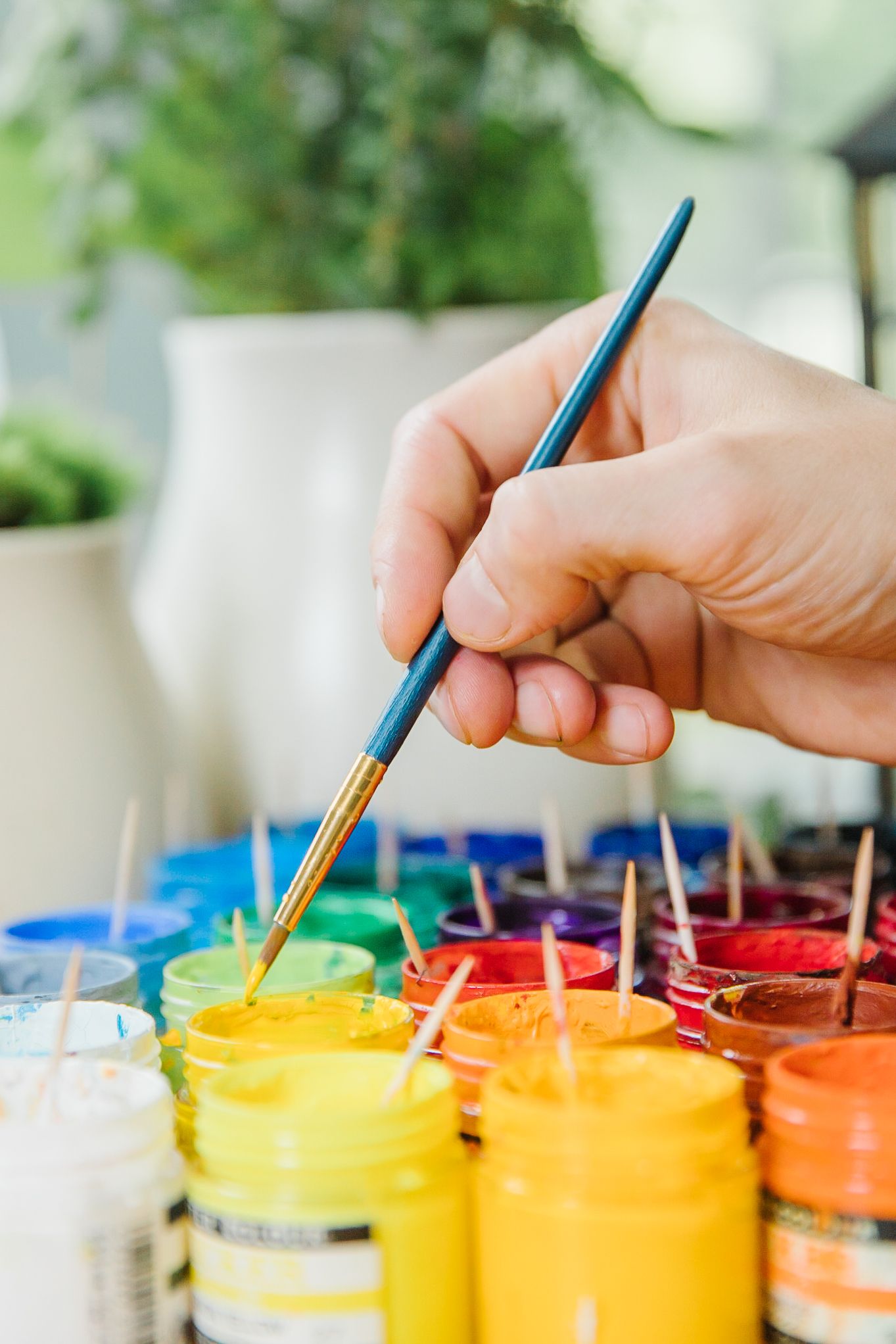 Hand holding blue paintbrush dipping into yellow paint with colorful paint jars lined up in the background.