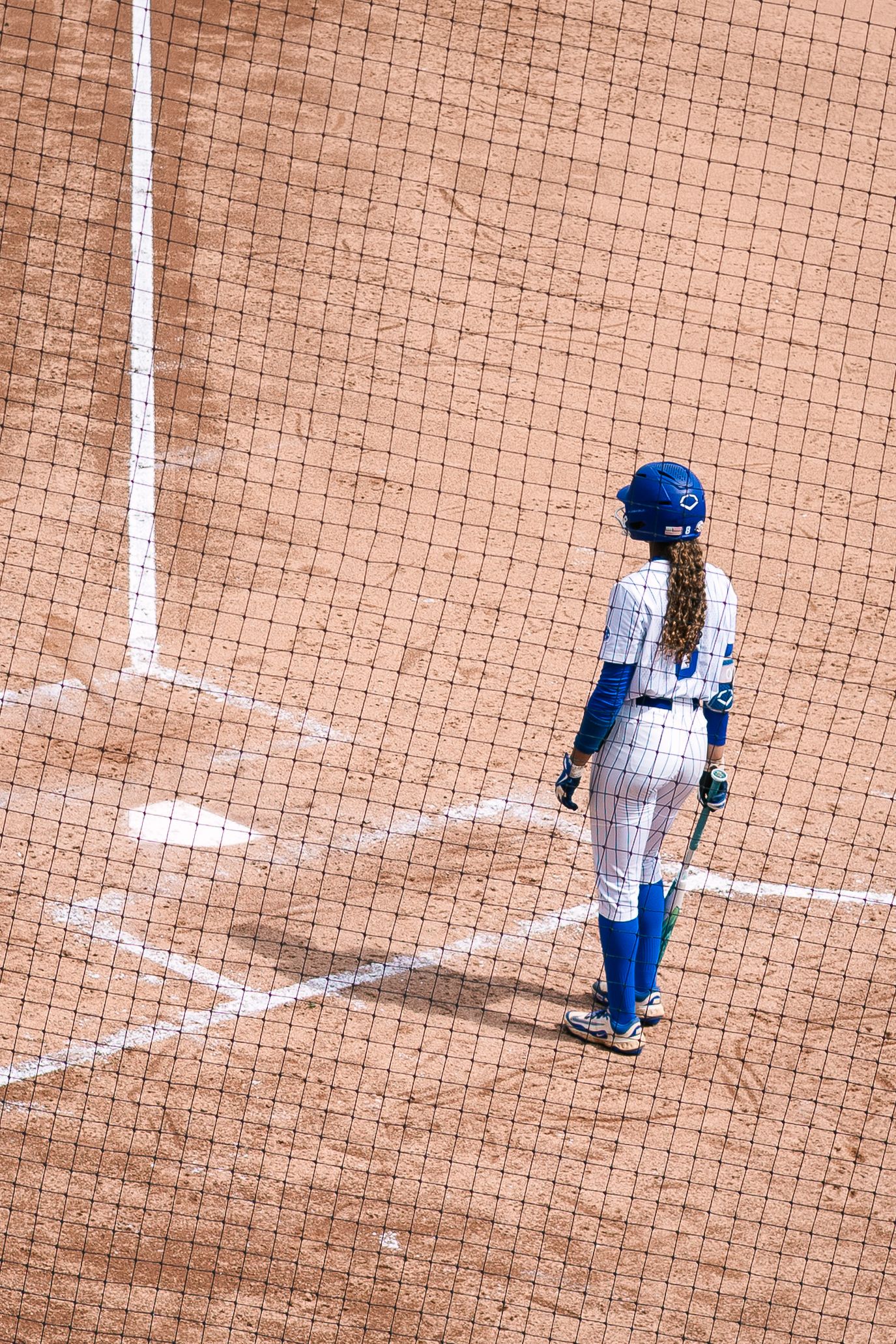 Female softball player in blue uniform and helmet stands at home plate on a dirt infield during a game.