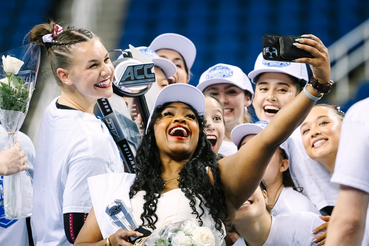 Excited fans in white baseball caps cheer and take selfies during a sporting event in a stadium.