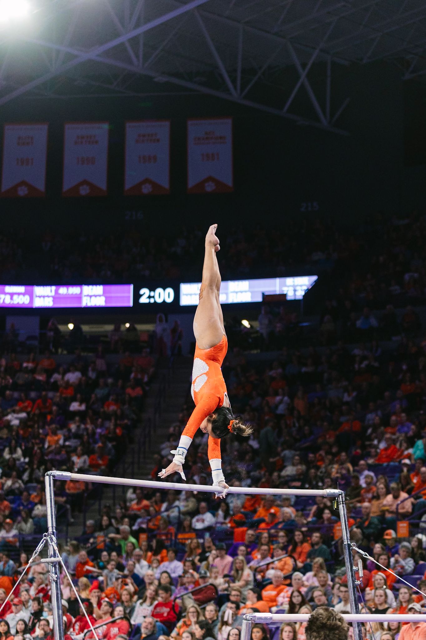 Gymnast performs a handstand on uneven bars in an orange leotard during a college gymnastics meet in a packed arena.