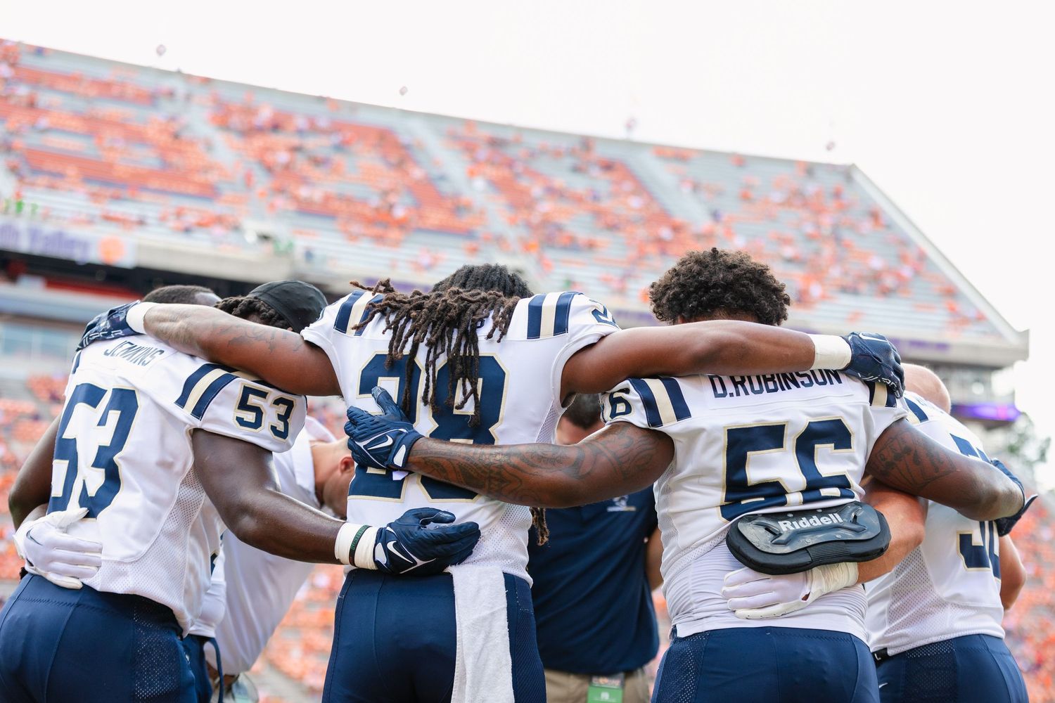 Football players in white uniforms huddle together on the field before a game in a packed stadium.