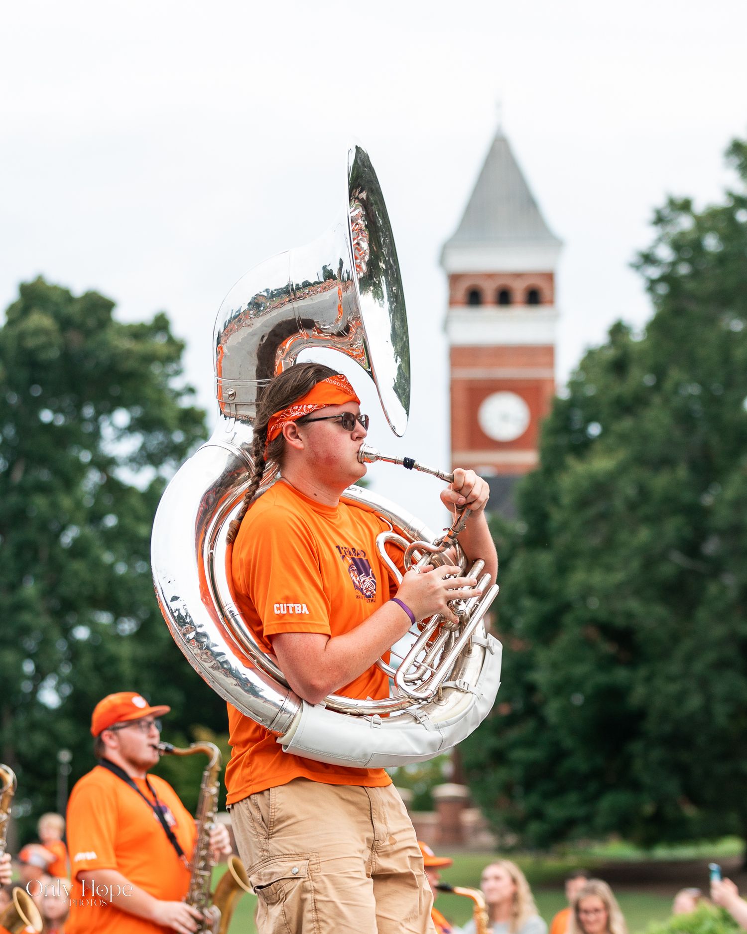 A sousaphone player in orange performs with a marching band in front of a brick tower with a clock.