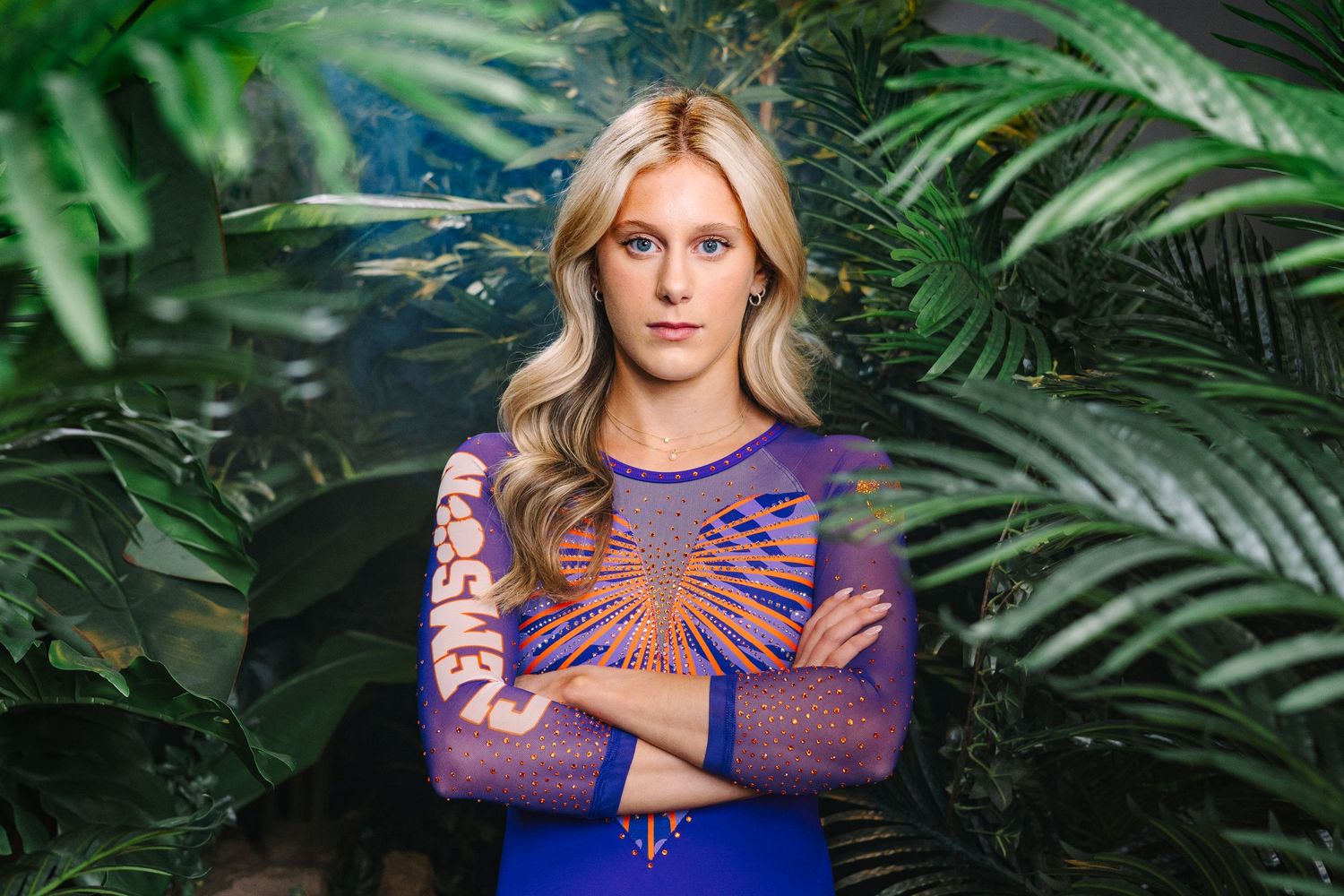 Model wearing a purple sheer embroidered bodysuit against lush tropical foliage background.