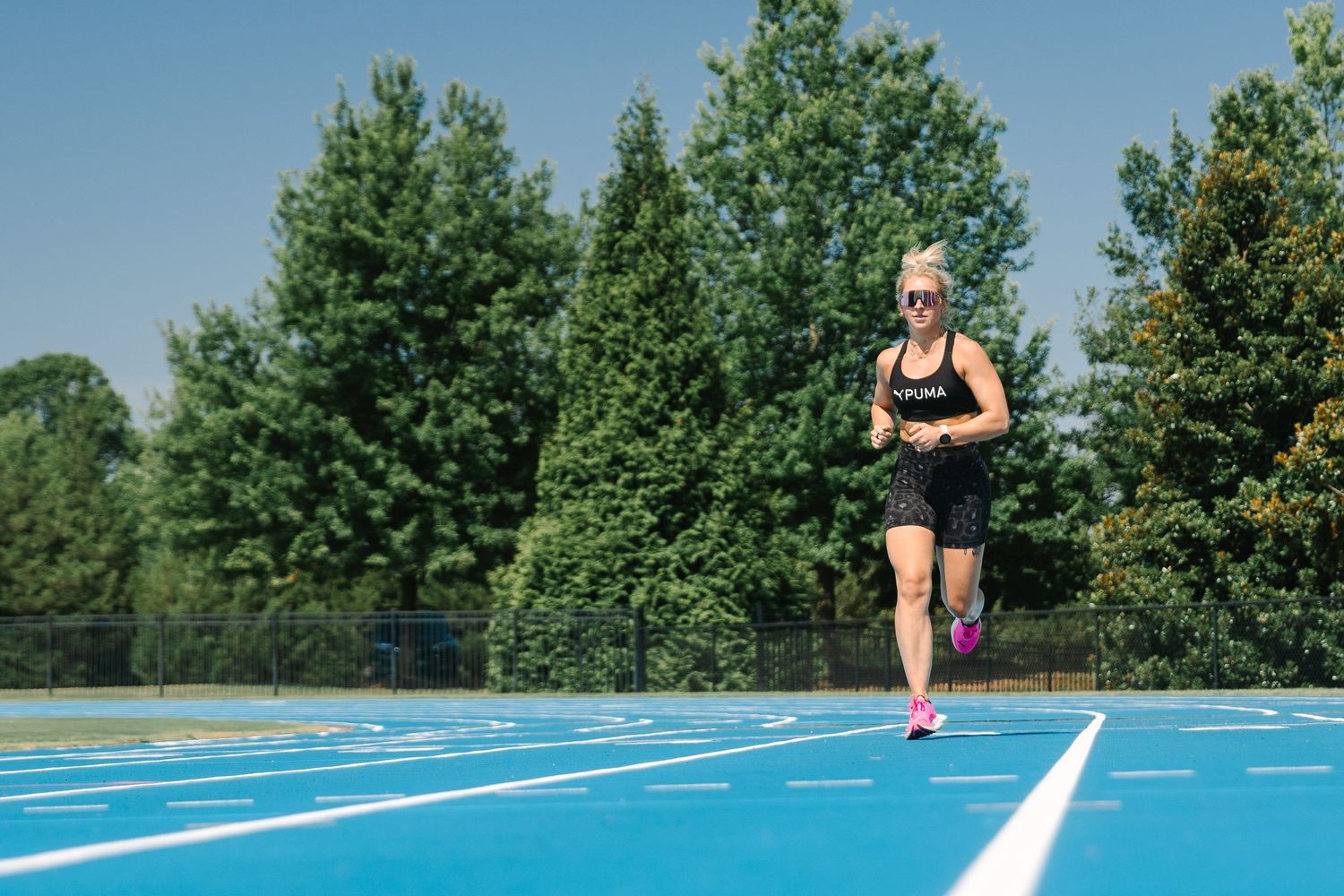 Woman in black athletic wear running on a blue outdoor track surrounded by green trees on a sunny day.