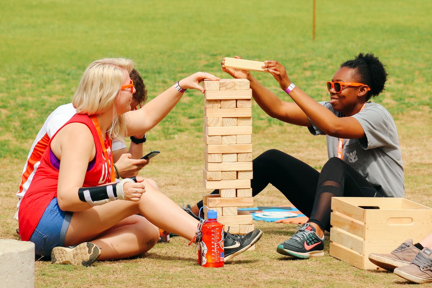 Two friends playing an outdoor game of Jenga on the grass with soda cans nearby, enjoying a sunny day.