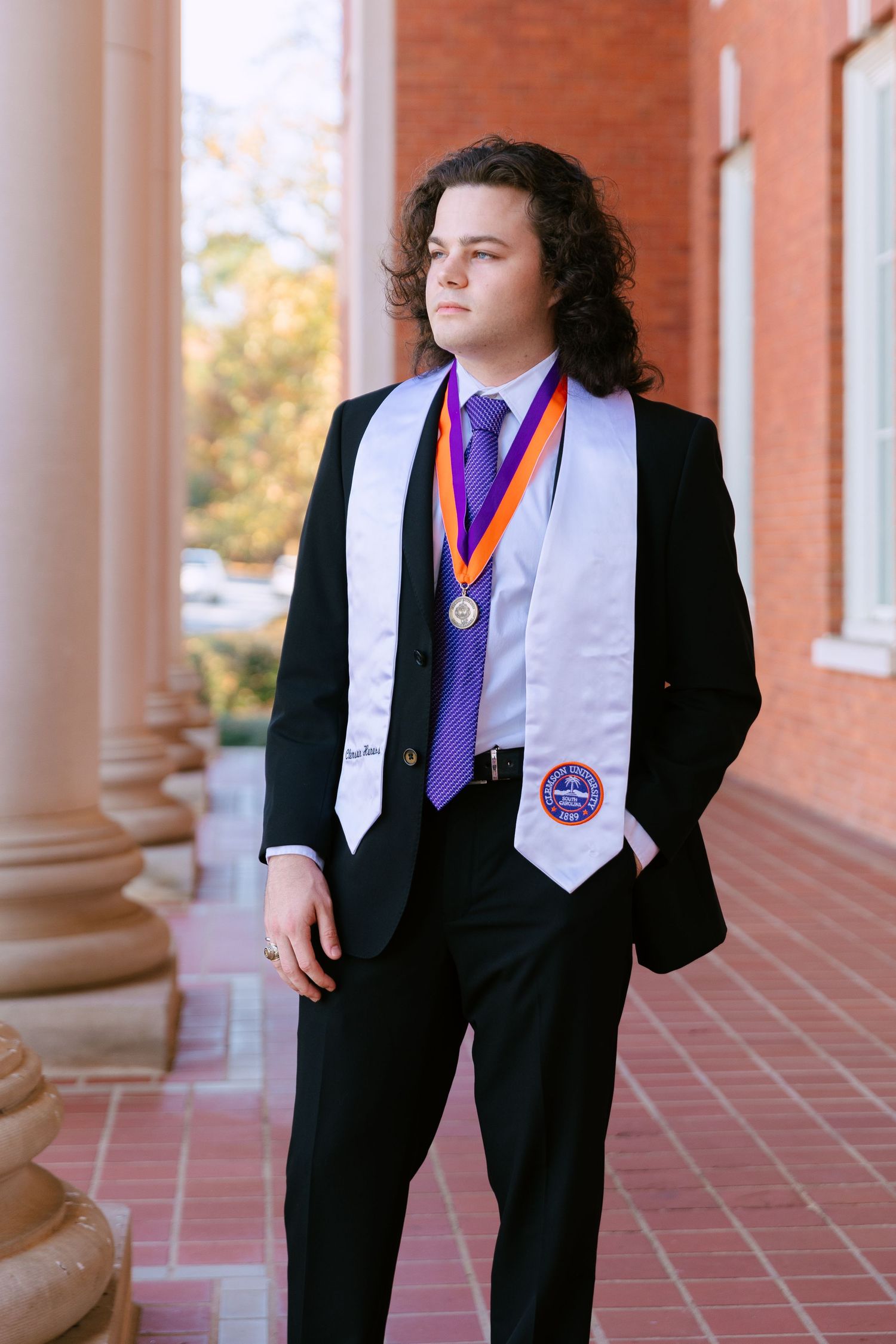 Young man in black suit with white graduation stole and medal poses outdoors near brick building columns.