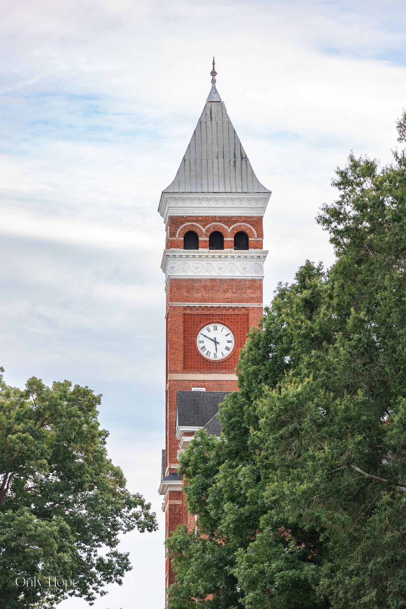 A historic red brick clock tower with white trim rises above green trees against a cloudy sky.