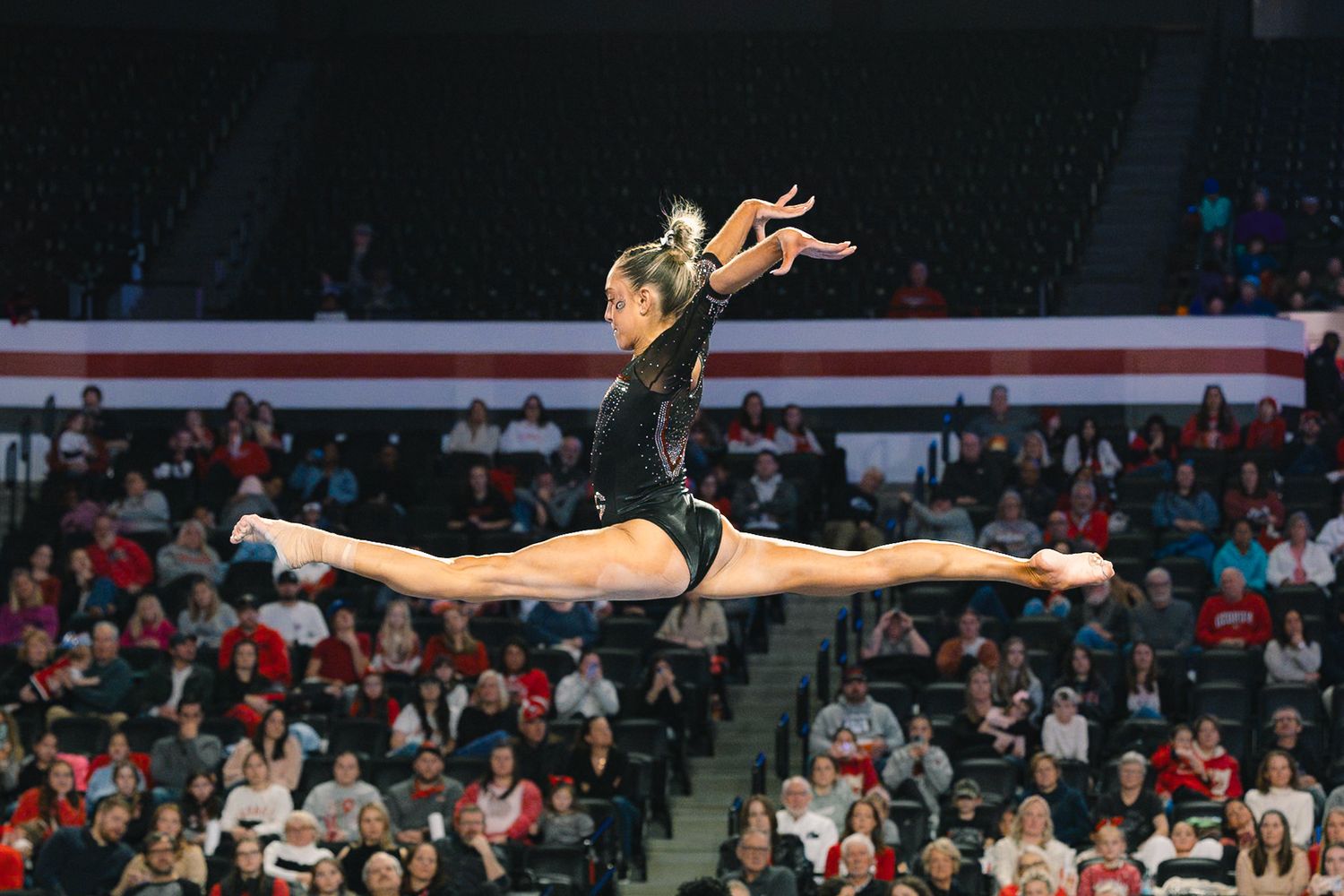 Gymnast performs a perfect split leap mid-air during a gymnastics competition in a packed arena.
