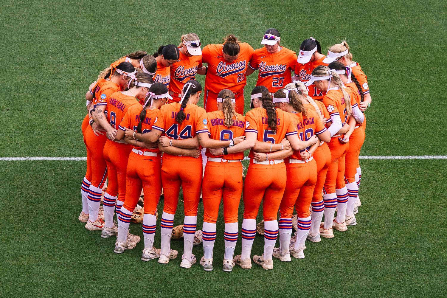 Clemson Tigers softball team huddles together in orange uniforms on a green field before a game.