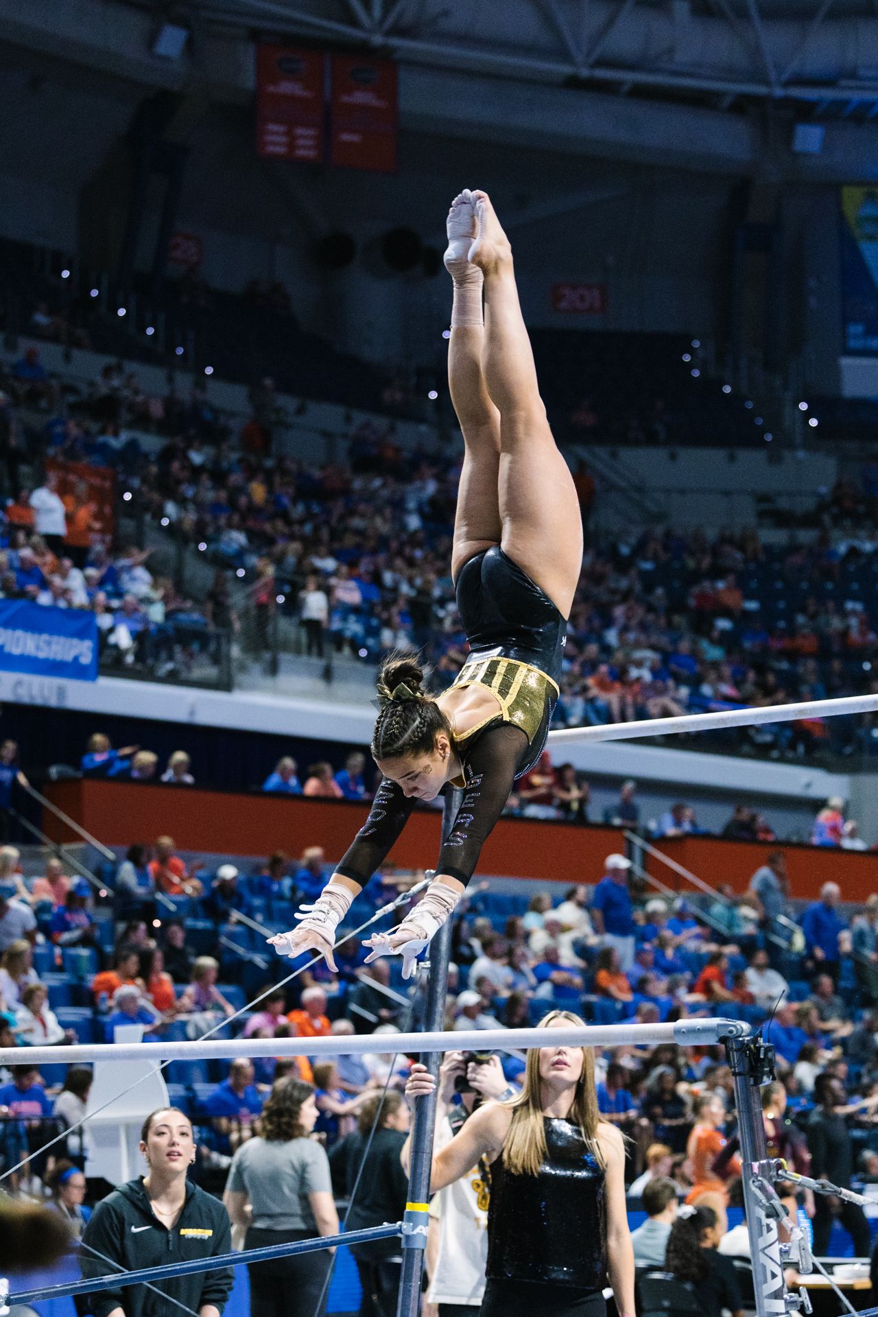 Gymnast performs a handstand on uneven bars during competitive gymnastics meet in arena with crowd watching.
