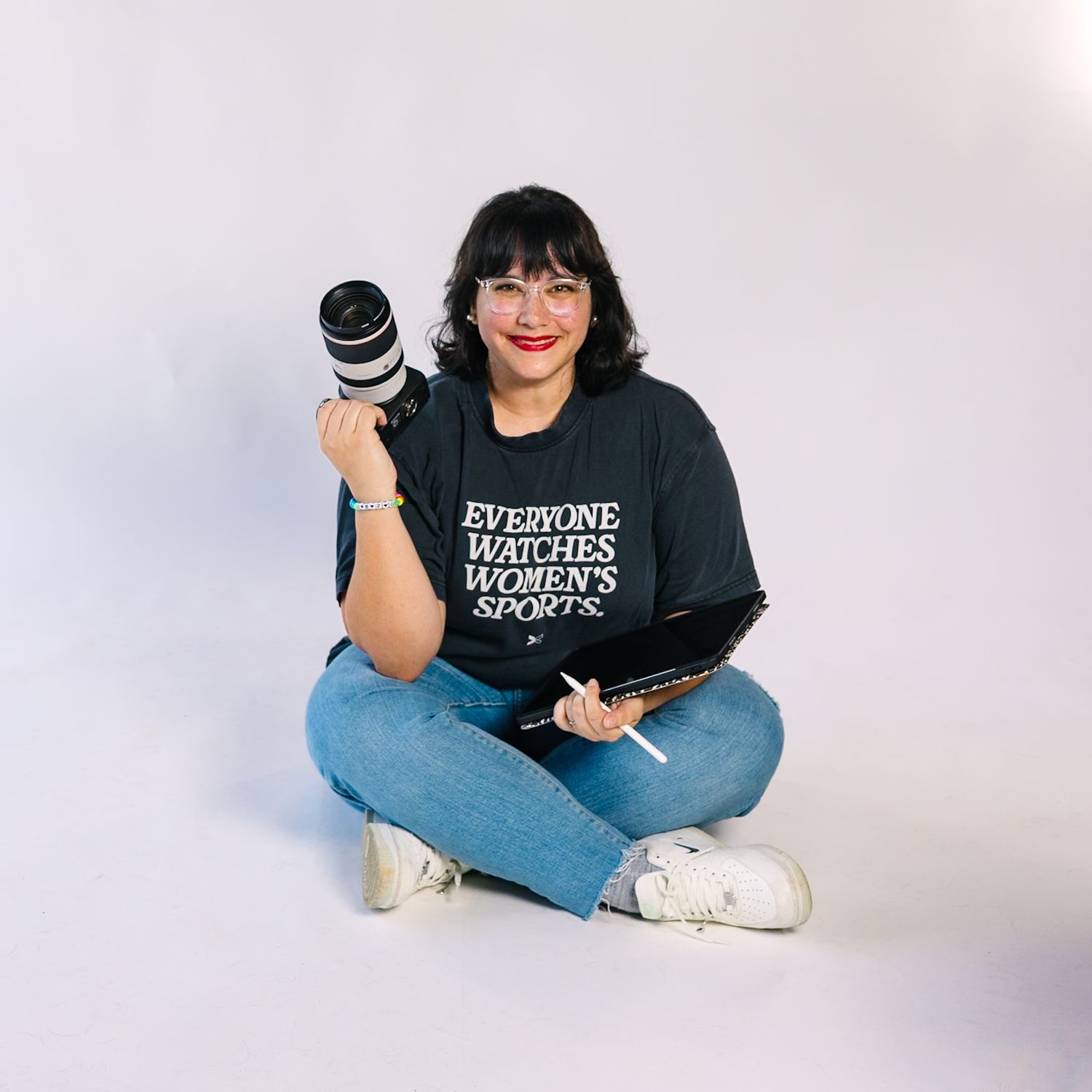 Photographer sits cross-legged on white background holding camera and laptop while wearing black t-shirt and jeans.