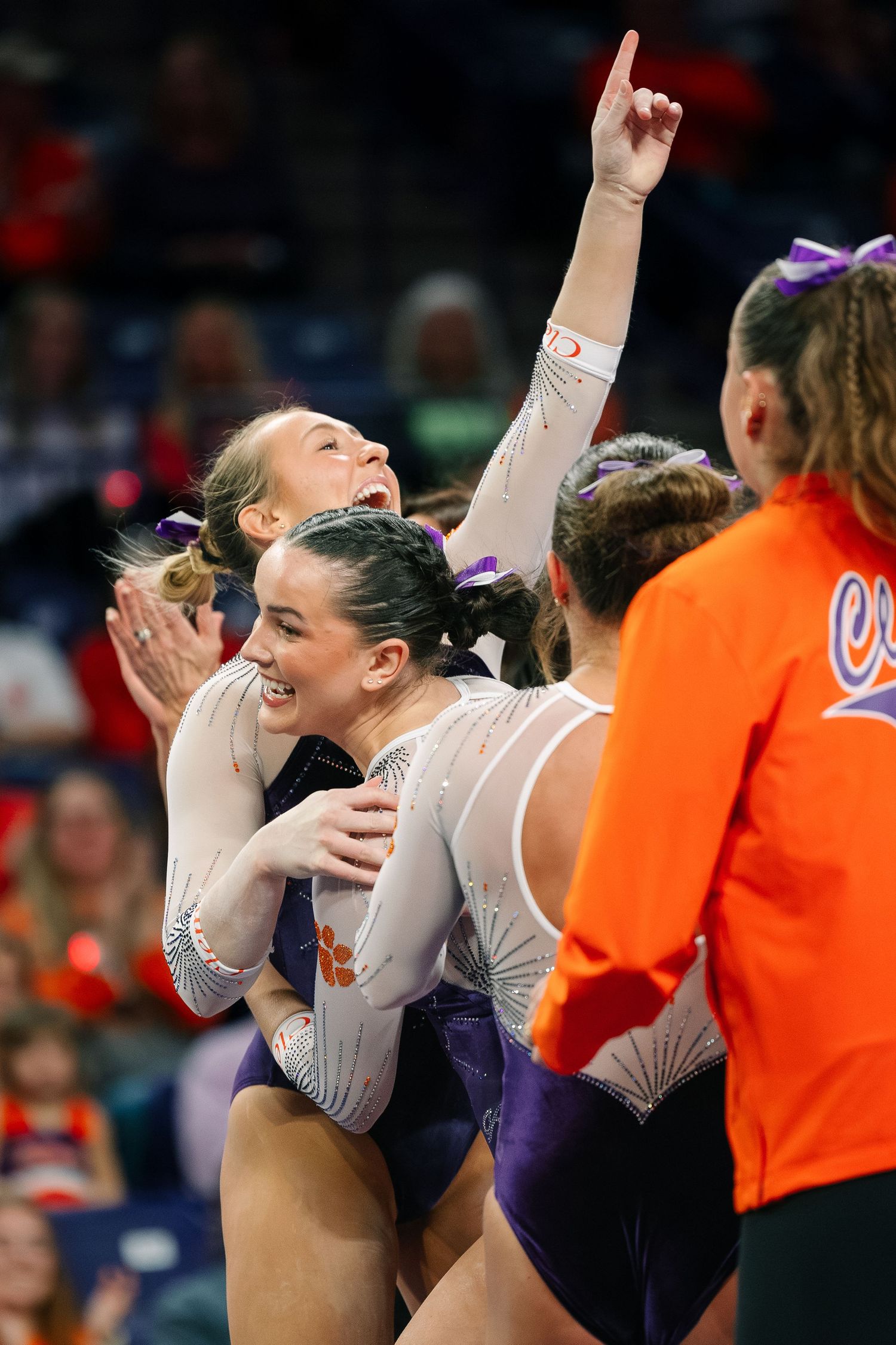 Clemson gymnasts celebrate together on the floor, wearing purple and orange leotards during a collegiate gymnastics meet.