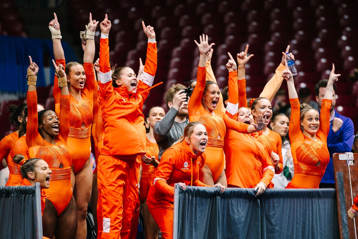 Enthusiastic team in bright orange uniforms cheering with raised arms from the sidelines at a sporting event.