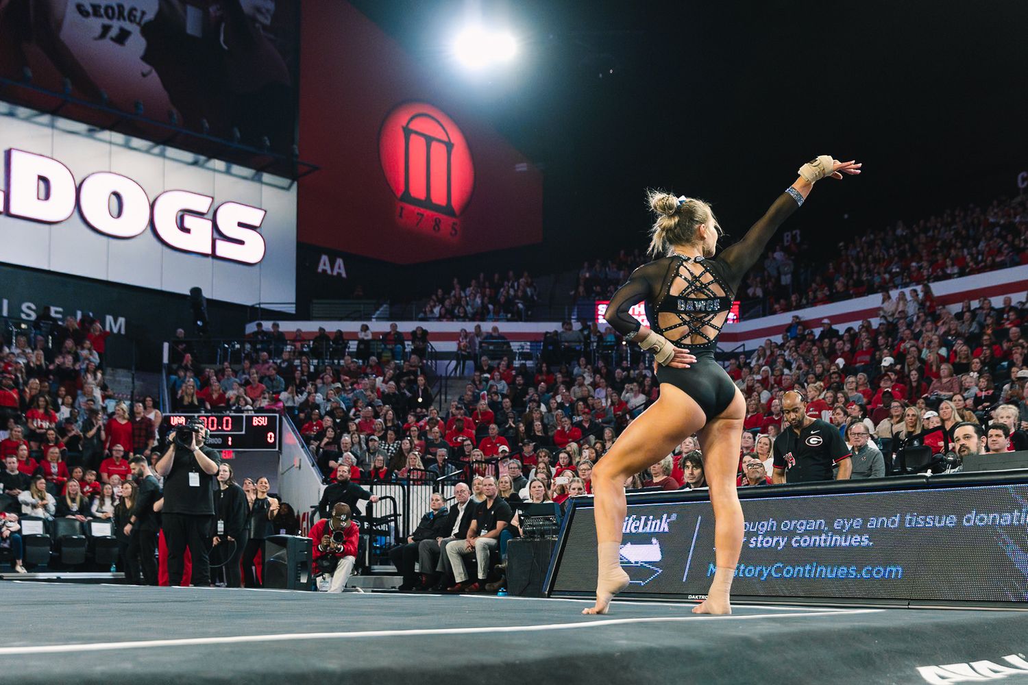 Gymnast performs dynamic floor routine during competition at packed indoor arena with bright spotlights illuminating performance.