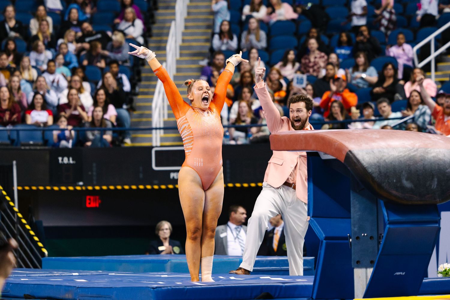 Gymnast in orange leotard performs on balance beam during collegiate competition.