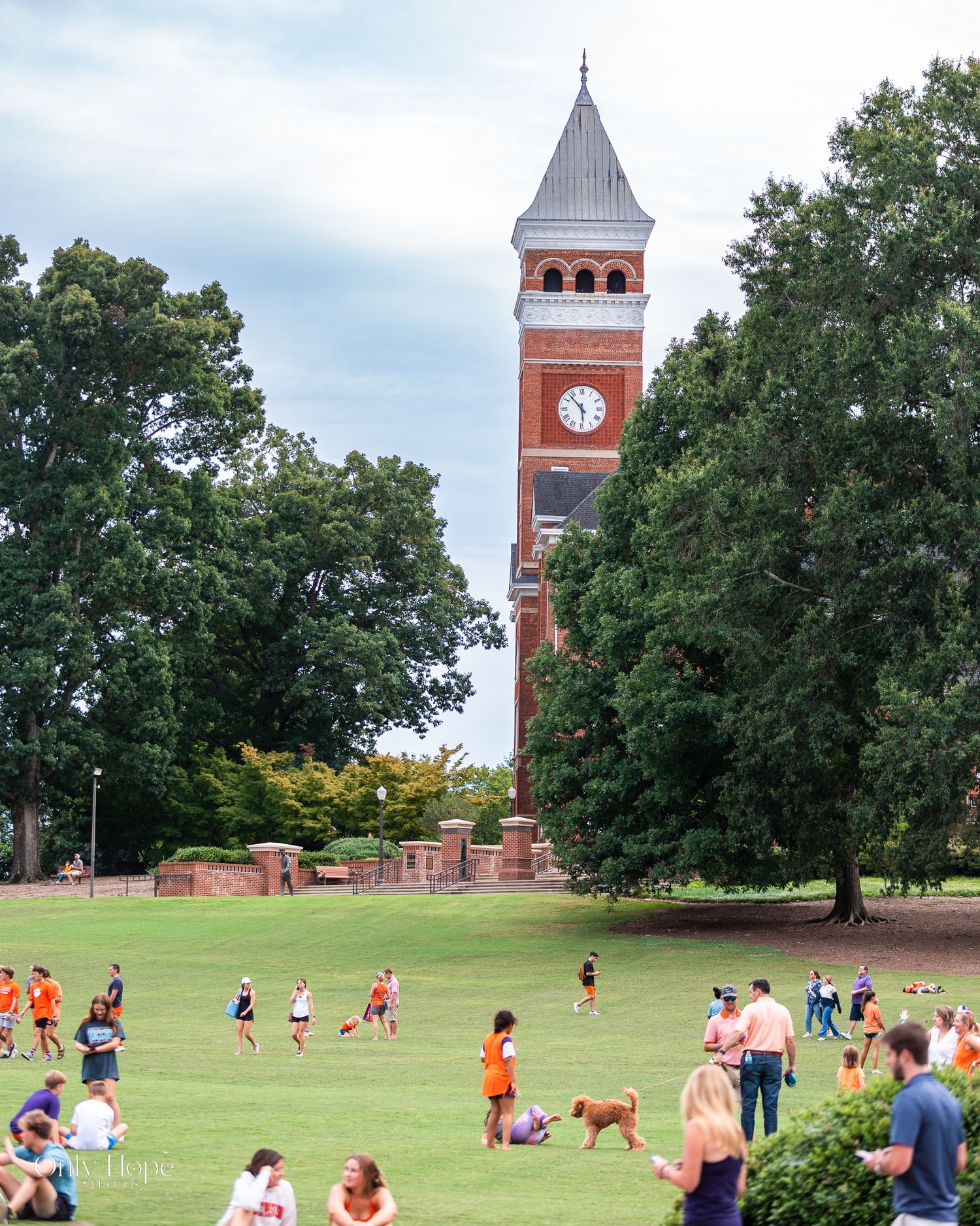 Students gather on a green campus lawn near a historic red brick clock tower at Clemson University on a cloudy day.