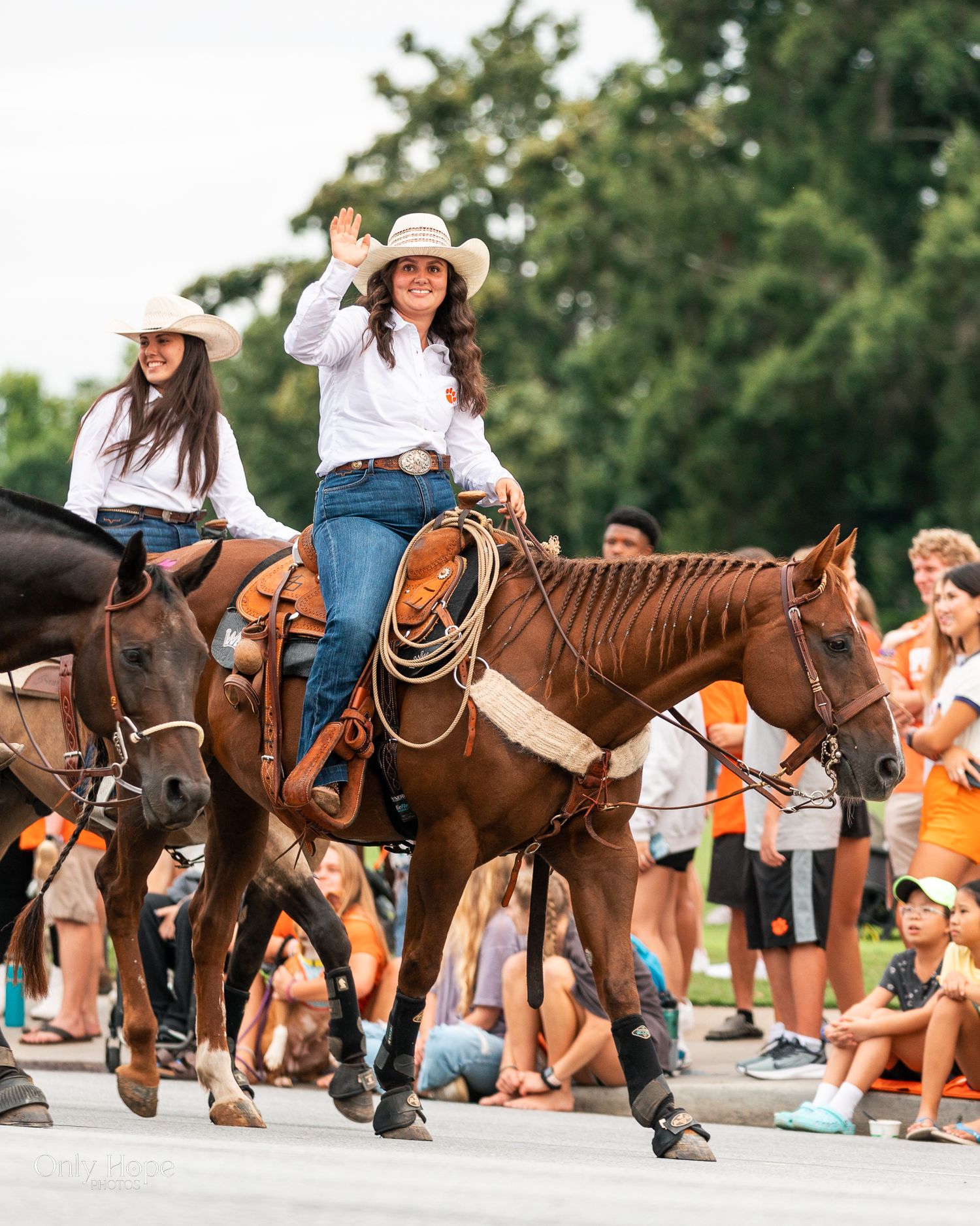 Riders on horseback wave to spectators during a parade as crowd watches from sidewalk on sunny summer day.