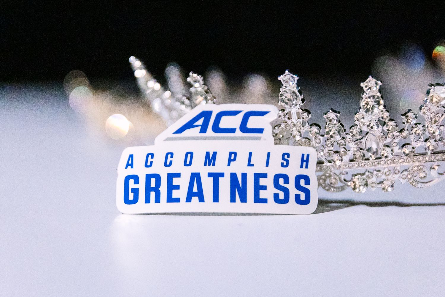 A decorative silver crown sits next to a blue ACC Accomplish Greatness logo in a shallow depth of field photo.