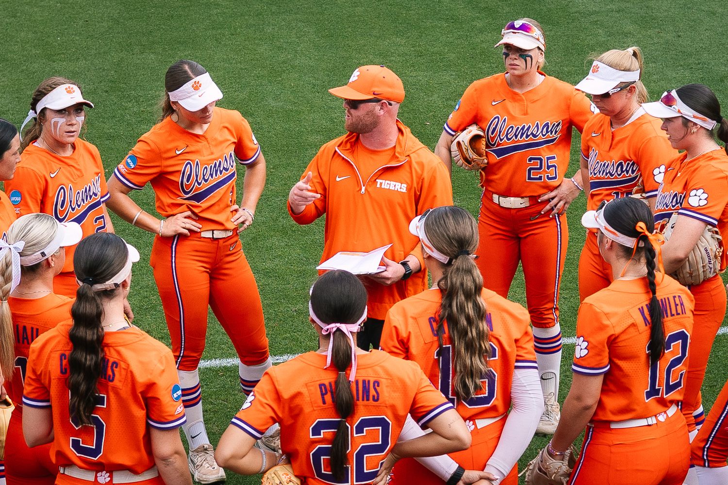 Clemson softball team huddles with coach in orange uniforms during a game discussion on the field.