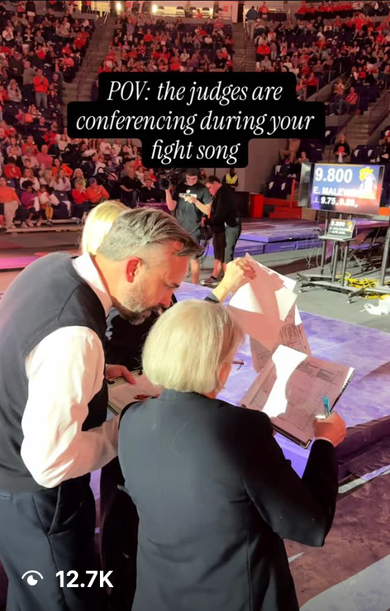 Judges conferencing over papers at a gymnastics competition with scores displayed on screen behind them.