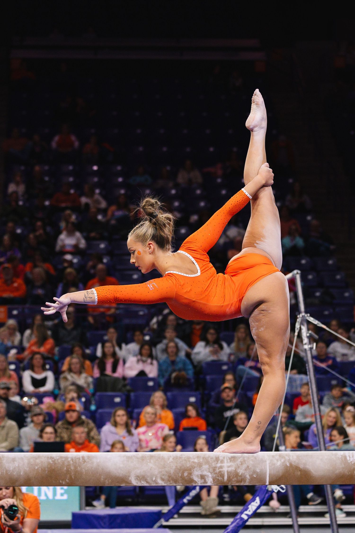Gymnast executing a skillful routine on the balance beam in an orange leotard.