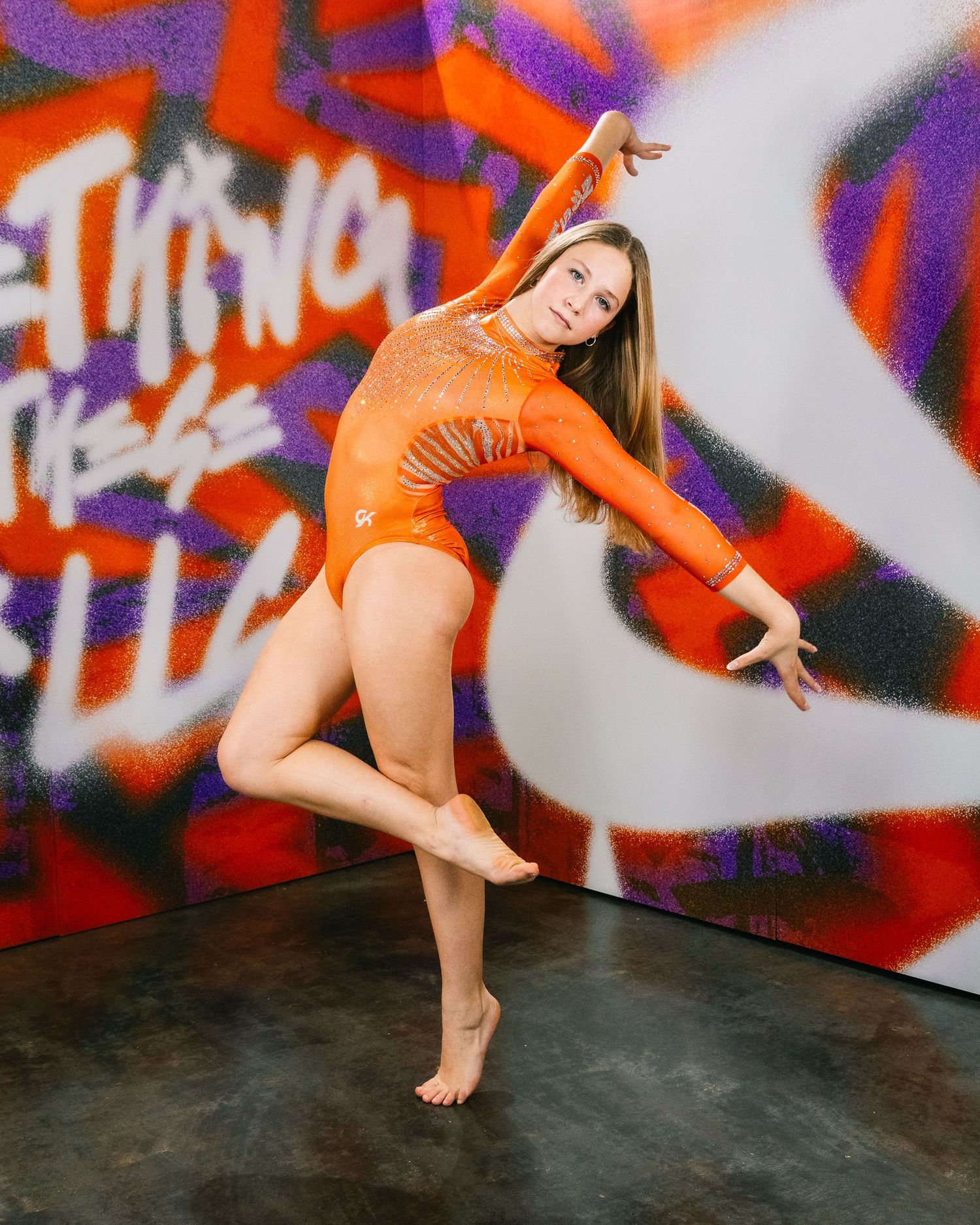 A dancer in an orange leotard poses expressively against a colorful graffiti wall.