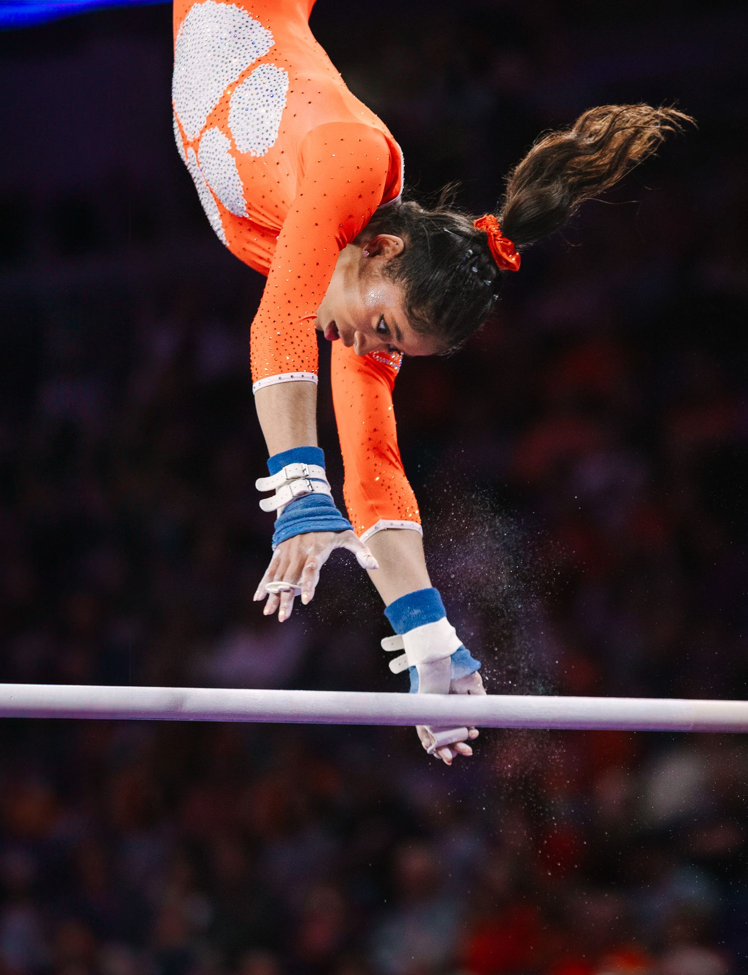 Gymnast in orange leotard performs dynamic uneven bars routine during competition.