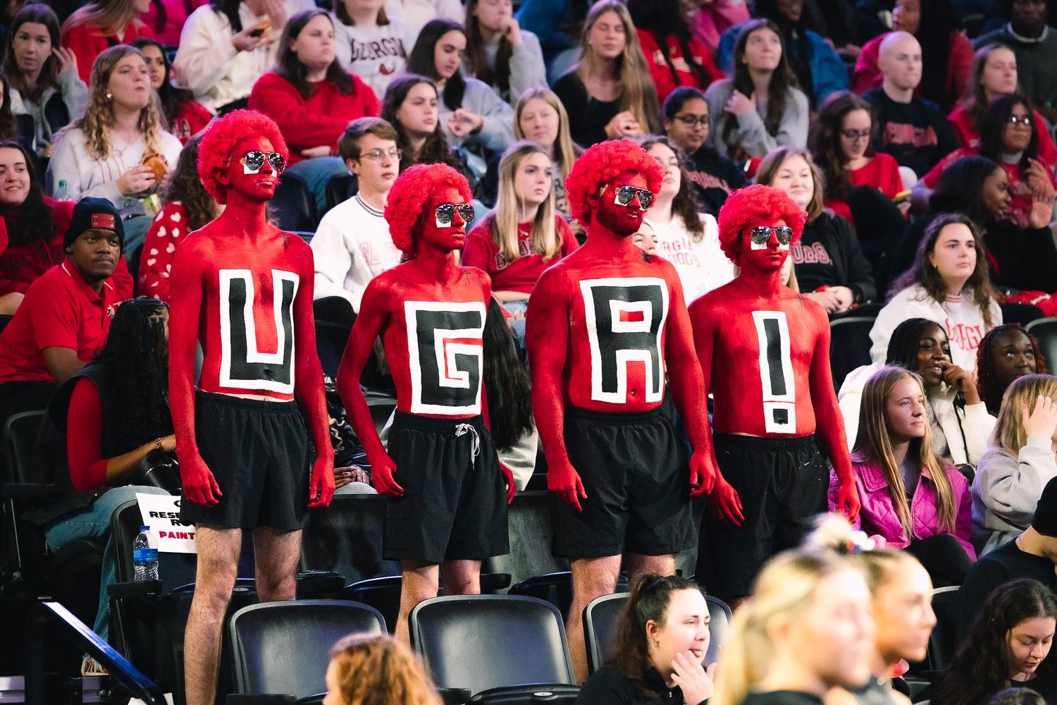 College sports fans wearing red jerseys with block letters stand in stadium bleachers as they cheer for their team.