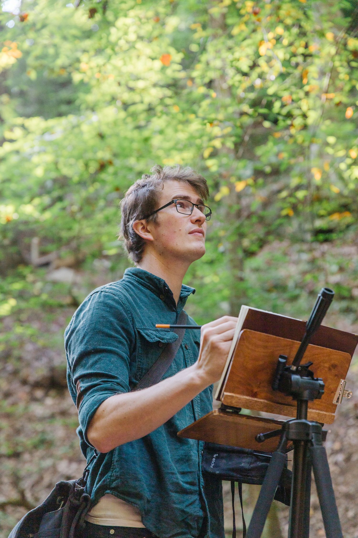 A photographer looks through a wooden view camera while shooting outdoors in a forest setting with green foliage.