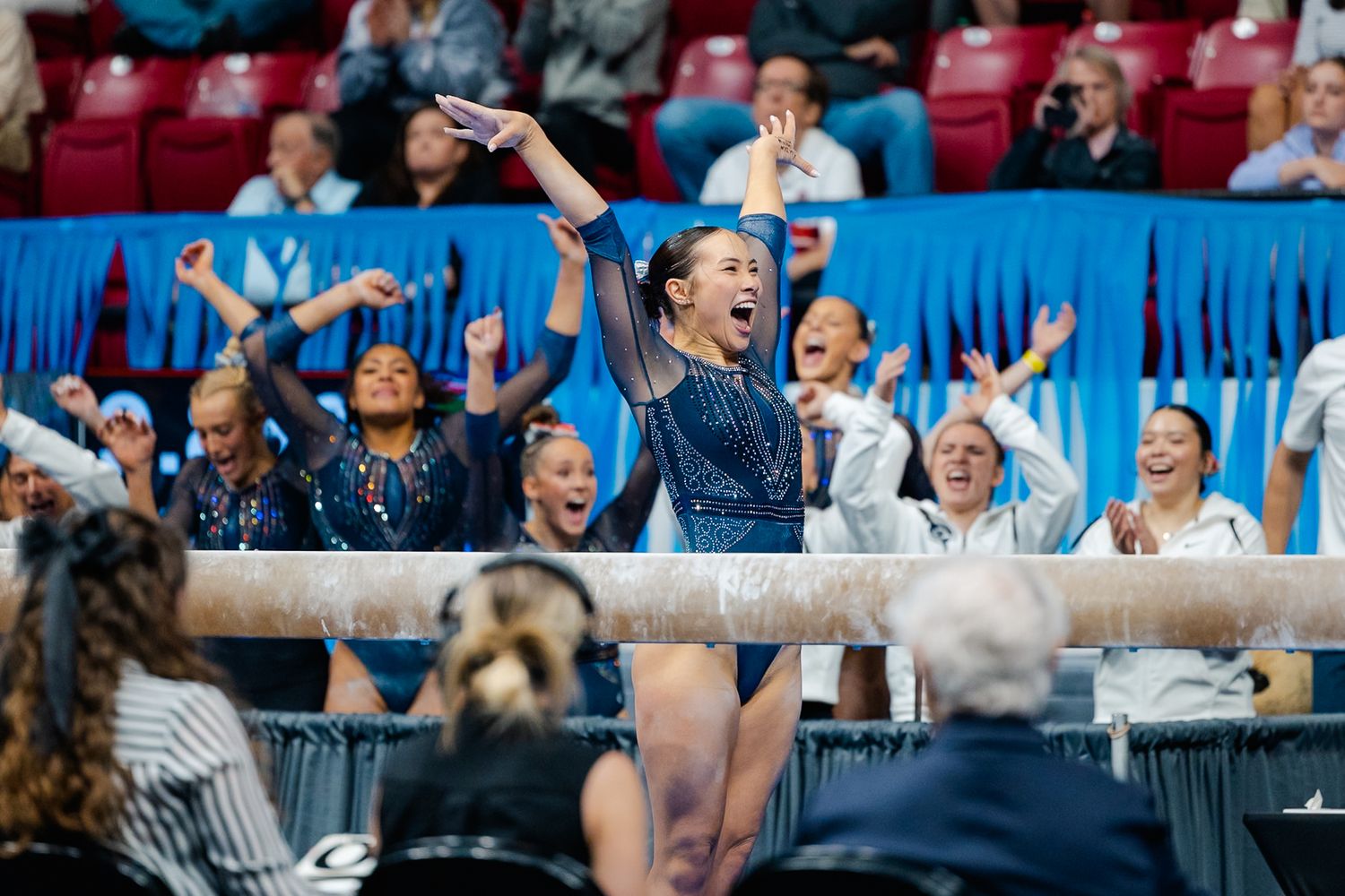 A gymnast performs on the uneven bars during a competition while teammates cheer enthusiastically in the background.