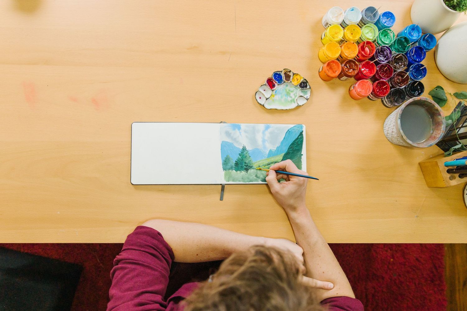 Artist using watercolor paints to create artwork on paper at wooden desk with colorful art supplies.