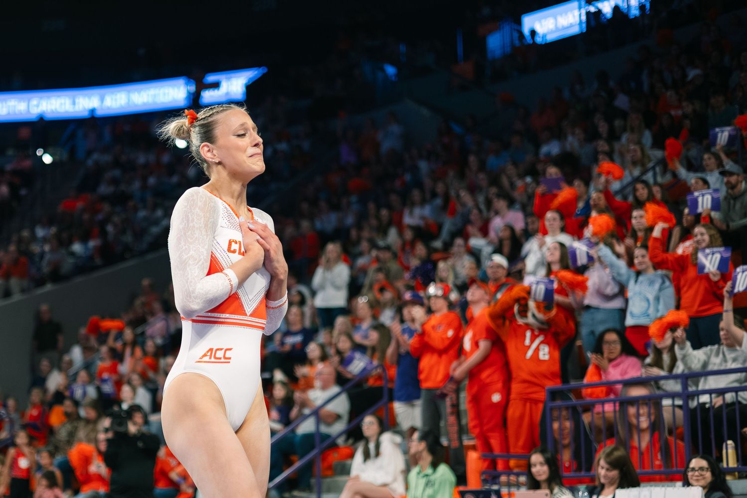 Female gymnast in orange ACC uniform stands poised before crowd at collegiate gymnastics competition.
