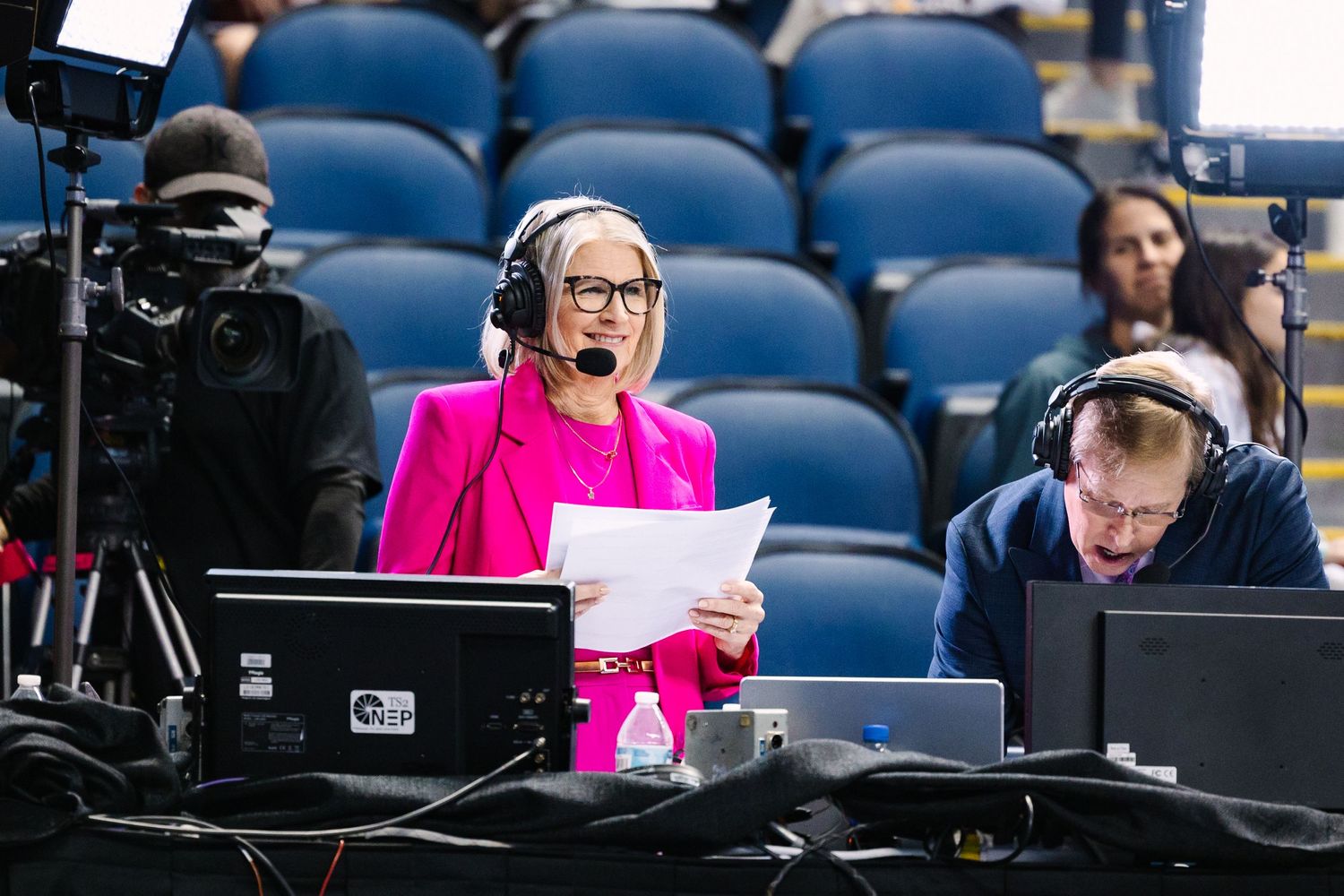 Commentator in bright pink blazer broadcasts from press box during sporting event.