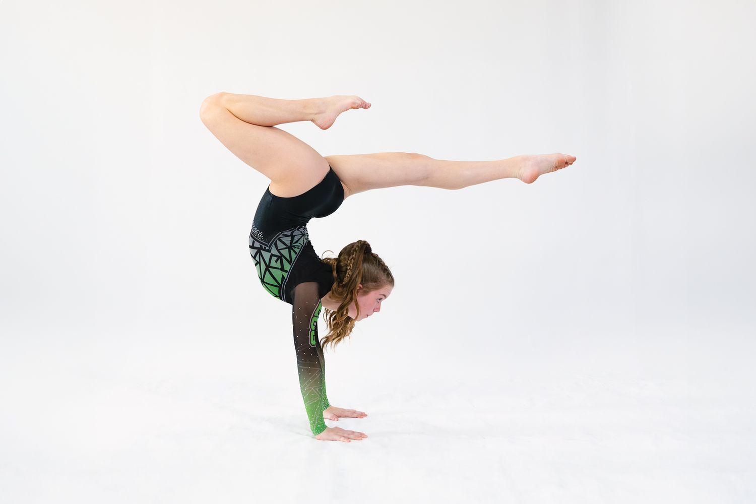 A gymnast performs a handstand split pose in a dark leotard against a bright white background.