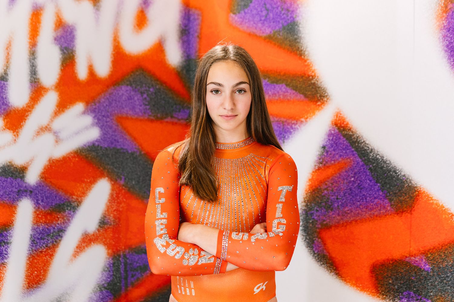Woman in bright orange sweater stands with crossed arms against colorful graffiti wall with red and purple spray paint.