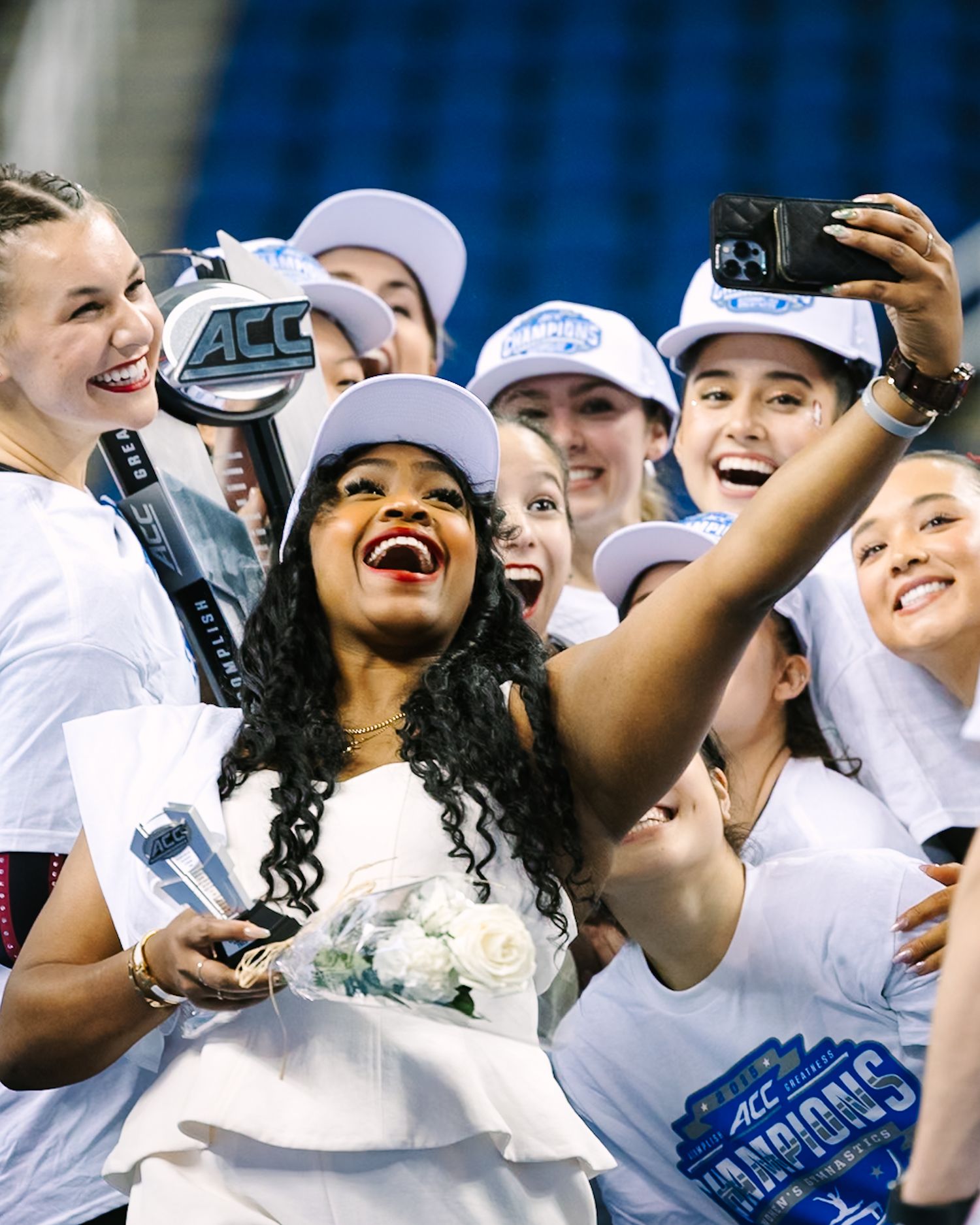 Women celebrating an ACC championship victory, taking a selfie with trophy, wearing white caps and shirts.