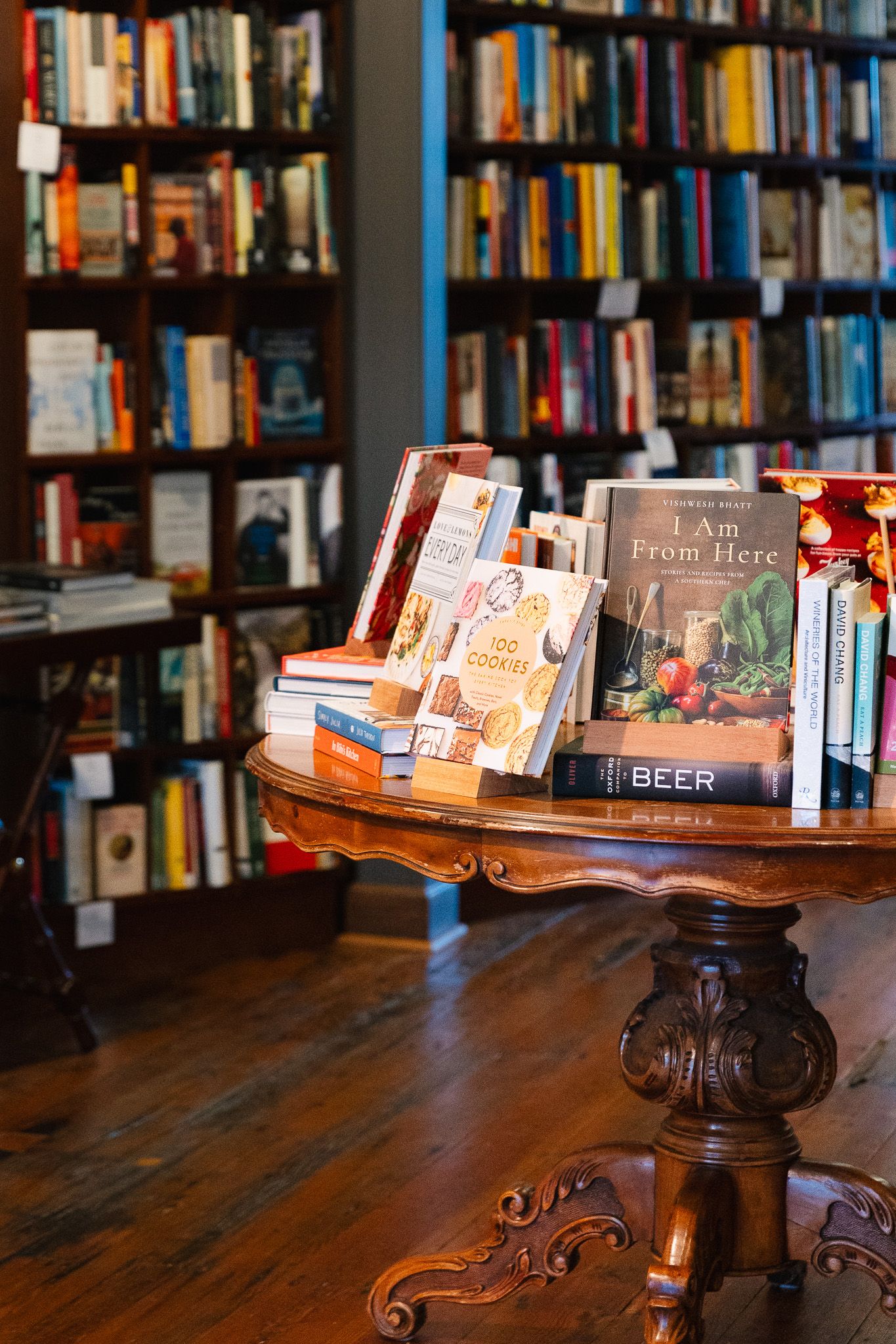 Books displayed on an antique carved wooden pedestal table with ornate details against a wall of filled bookshelves.