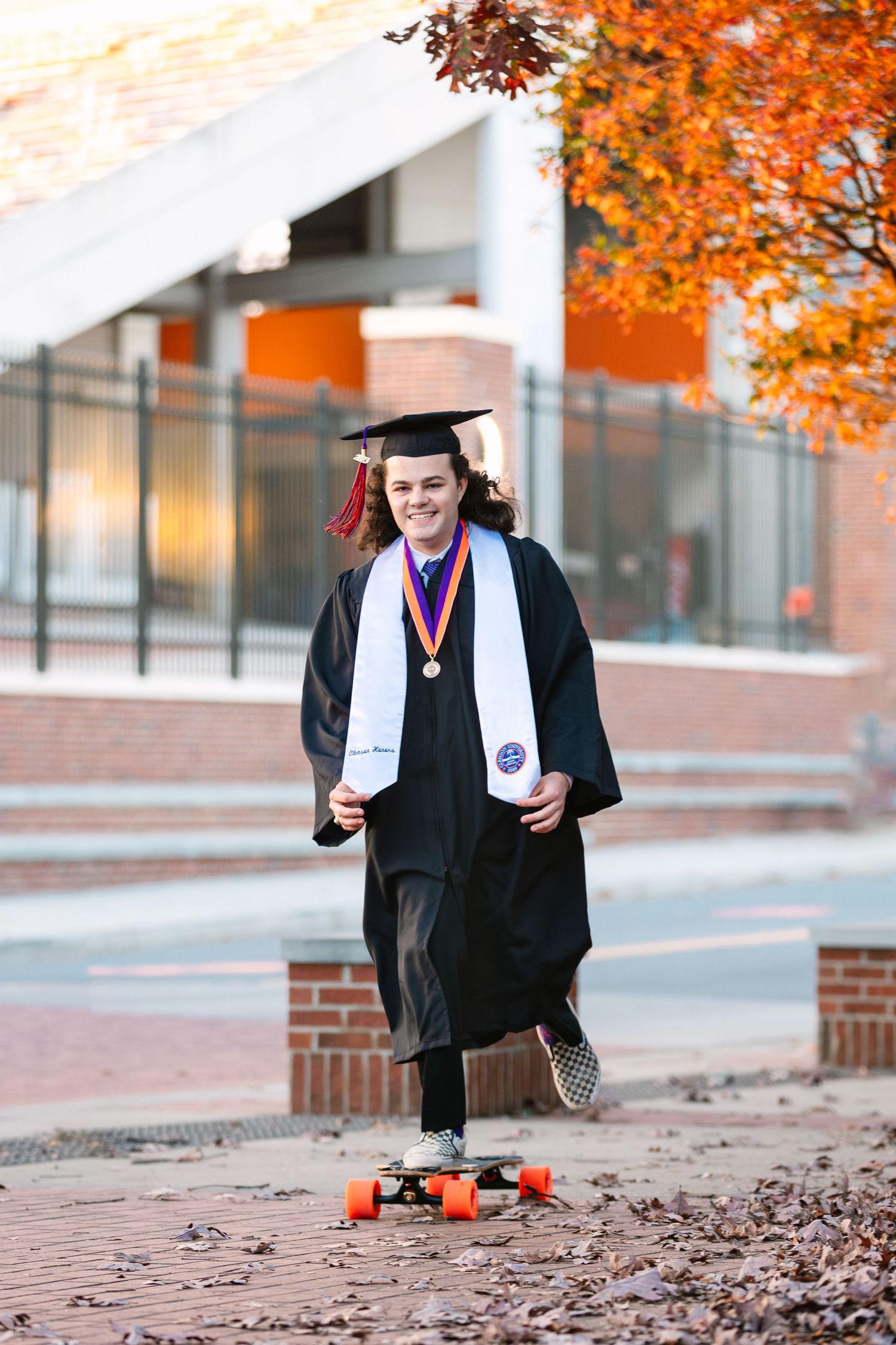 Graduate in cap and gown rides a skateboard on campus during fall, wearing medals and a white stole.