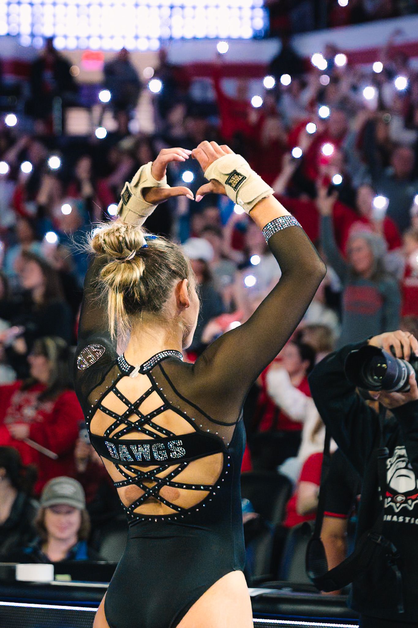 Gymnast in a black leotard with strappy back design performs during a competition in an arena with crowd.