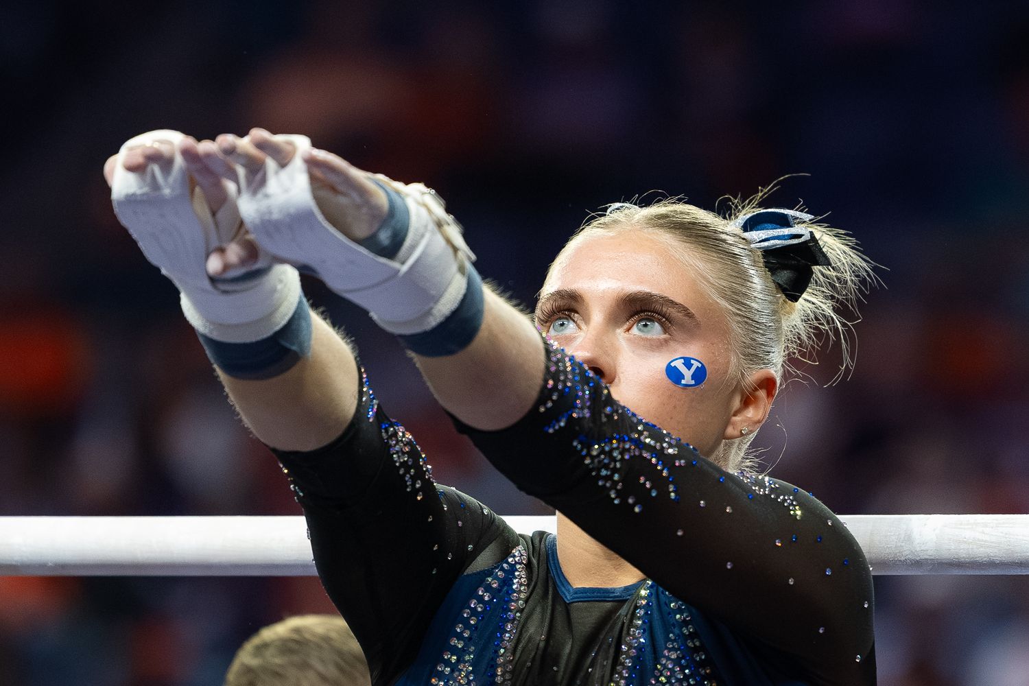 BYU gymnast in black leotard with blue face sticker chalking hands before competition on uneven bars.