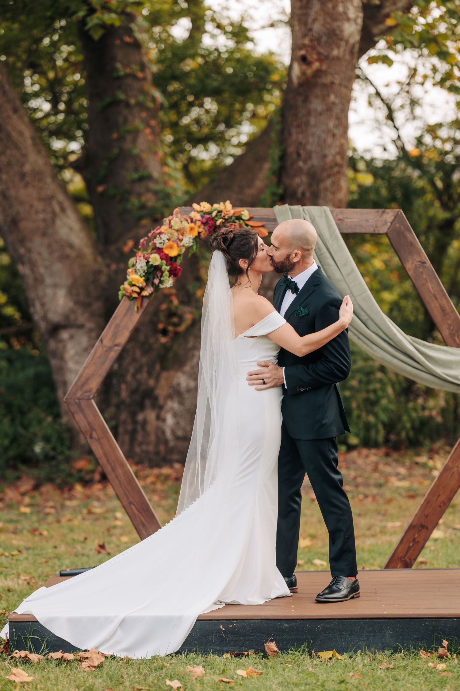 Wedding couple sharing romantic moment on wooden deck with draped fabric arch in autumn setting.