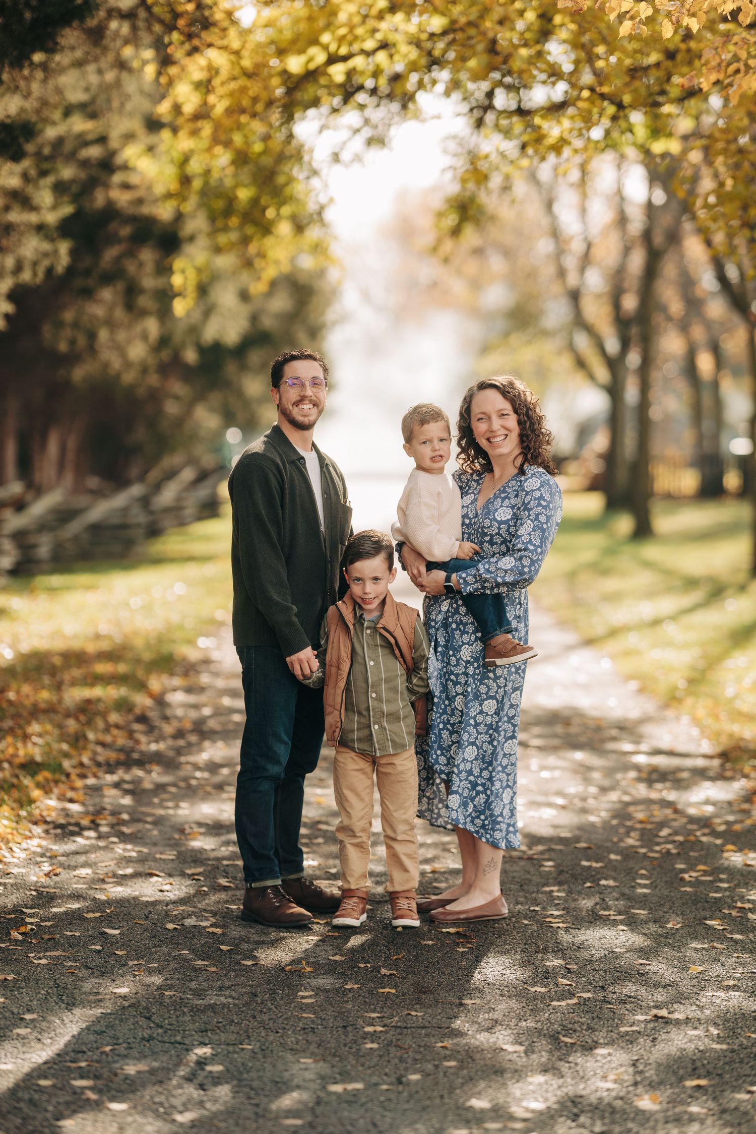 Family portrait session on a sunlit autumn path with golden leaves and trees creating a warm natural backdrop.