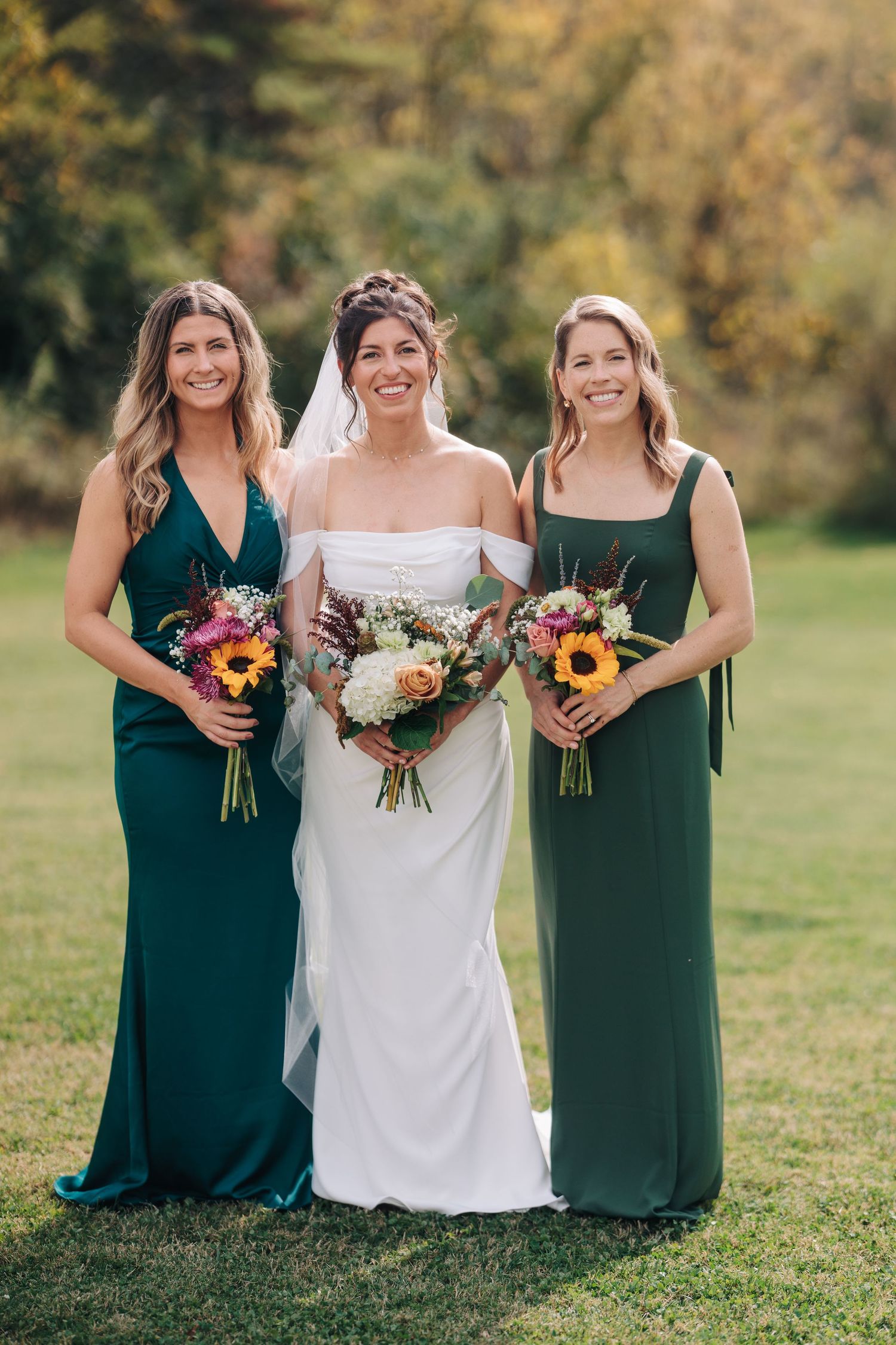 Wedding party in emerald green dresses holding colorful autumn bouquets on grassy field.