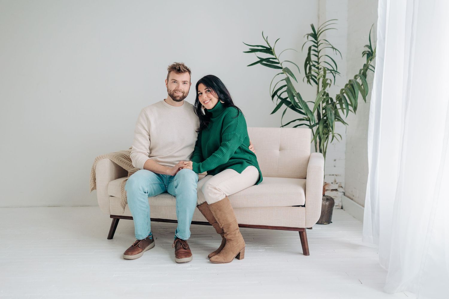 A couple sits close together on a beige couch in a minimalist white room with a potted palm plant.