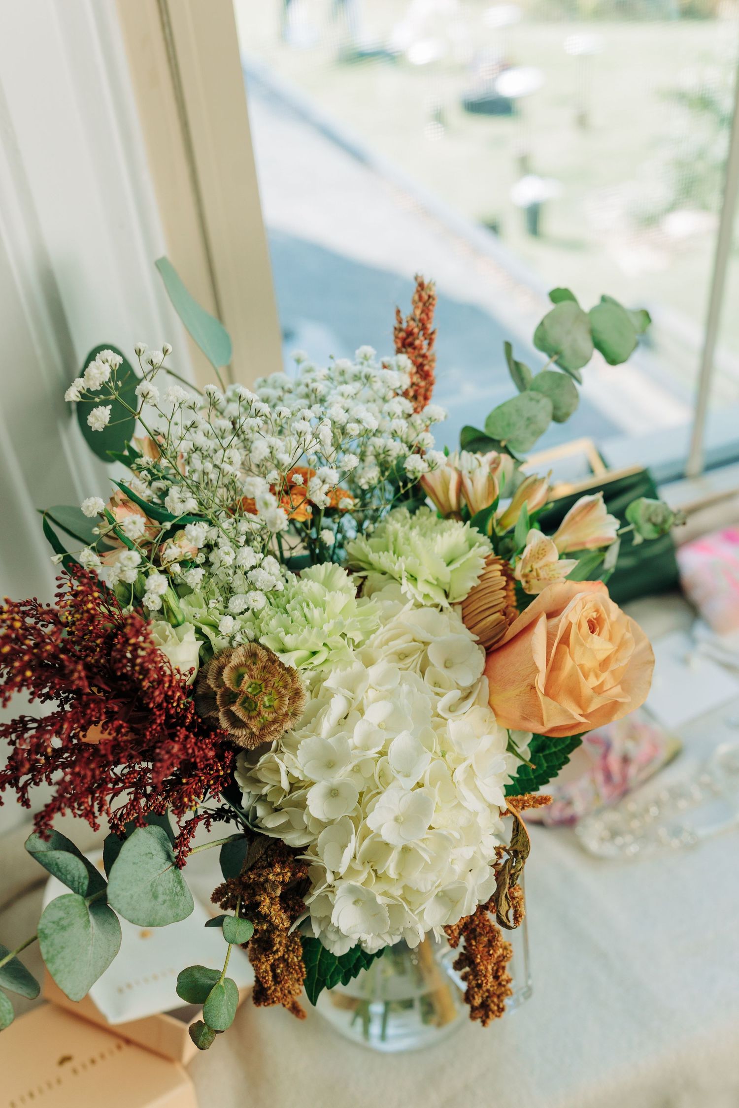 A romantic autumn wedding bouquet with white hydrangeas, peach roses, and eucalyptus arranged in a glass vase.