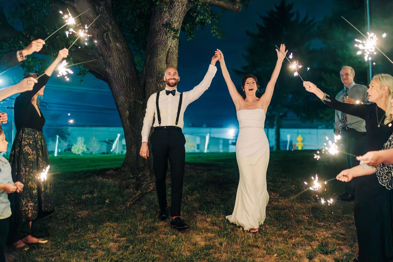 Wedding couple holds hands triumphantly under sparklers at nighttime outdoor celebration.
