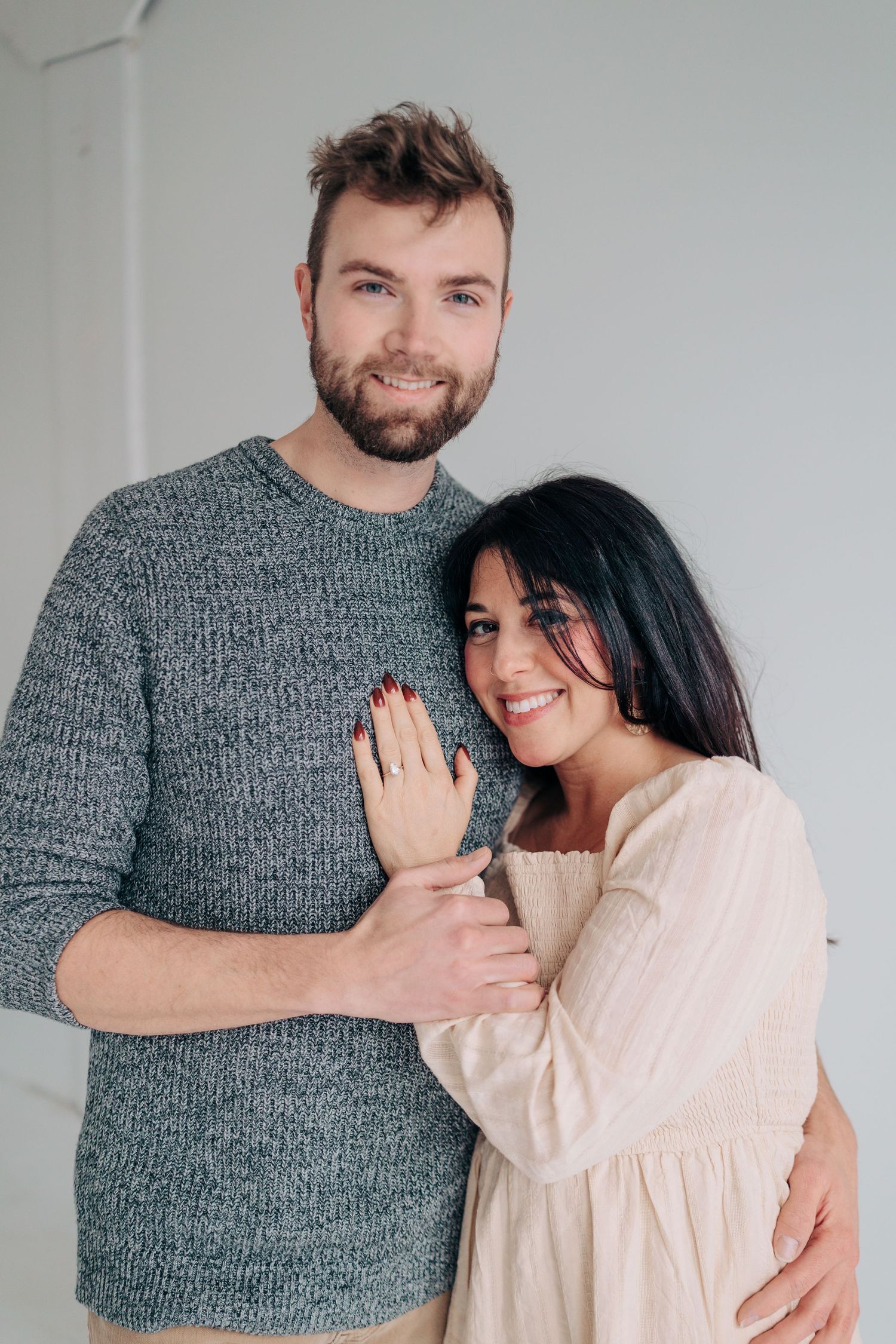 A couple in casual attire share tender moments in a series of romantic engagement photos against a white wall.