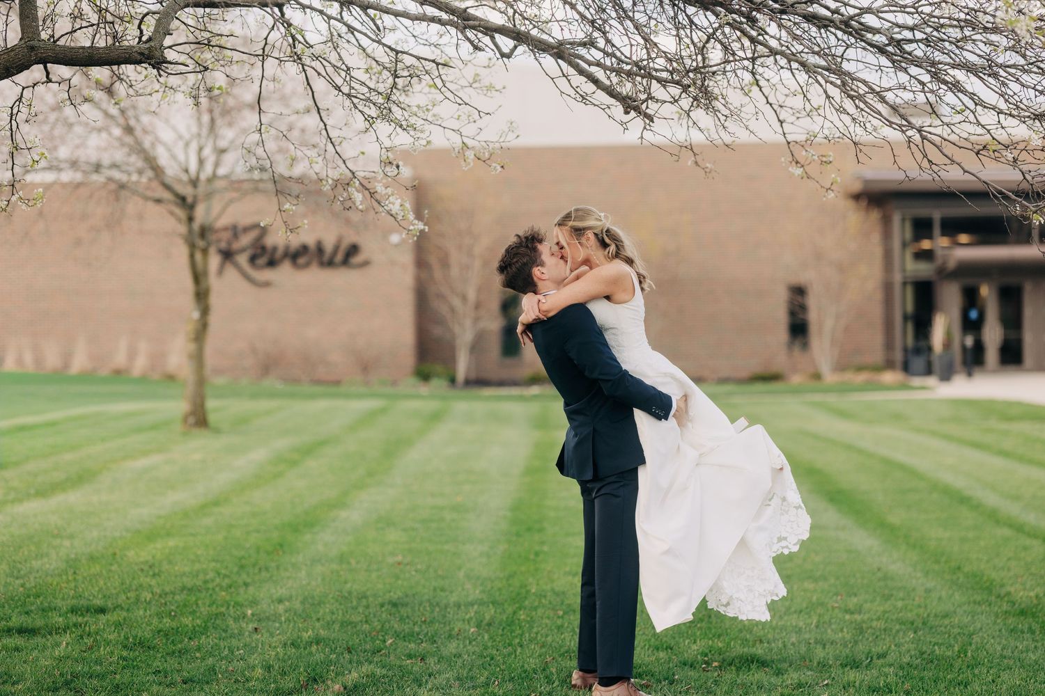 Groom lifting bride in wedding dress outside Reverie venue on lush green lawn.